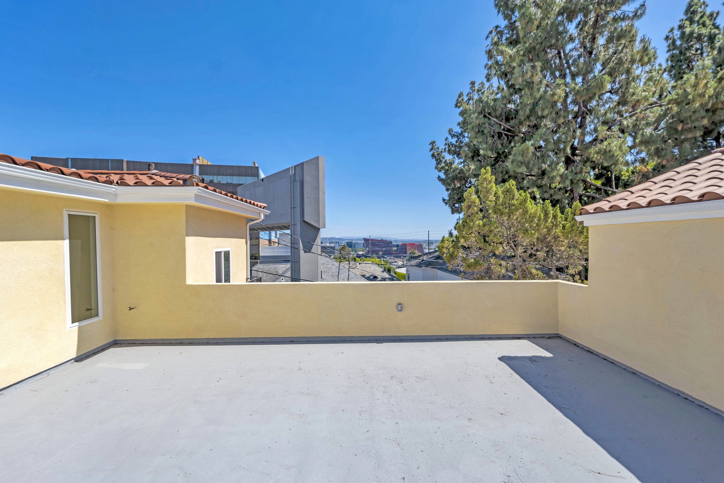 An empty outdoor patio with a concrete floor and a yellow wall, overlooking rooftops, trees, and a cityscape in the background under a clear blue sky.