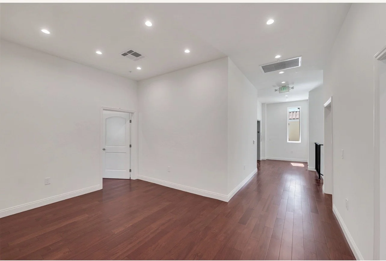 Empty room with white walls, hardwood floors, ceiling lights, and a door on the left. There is a window at the back and a small area with a black railing on the right.