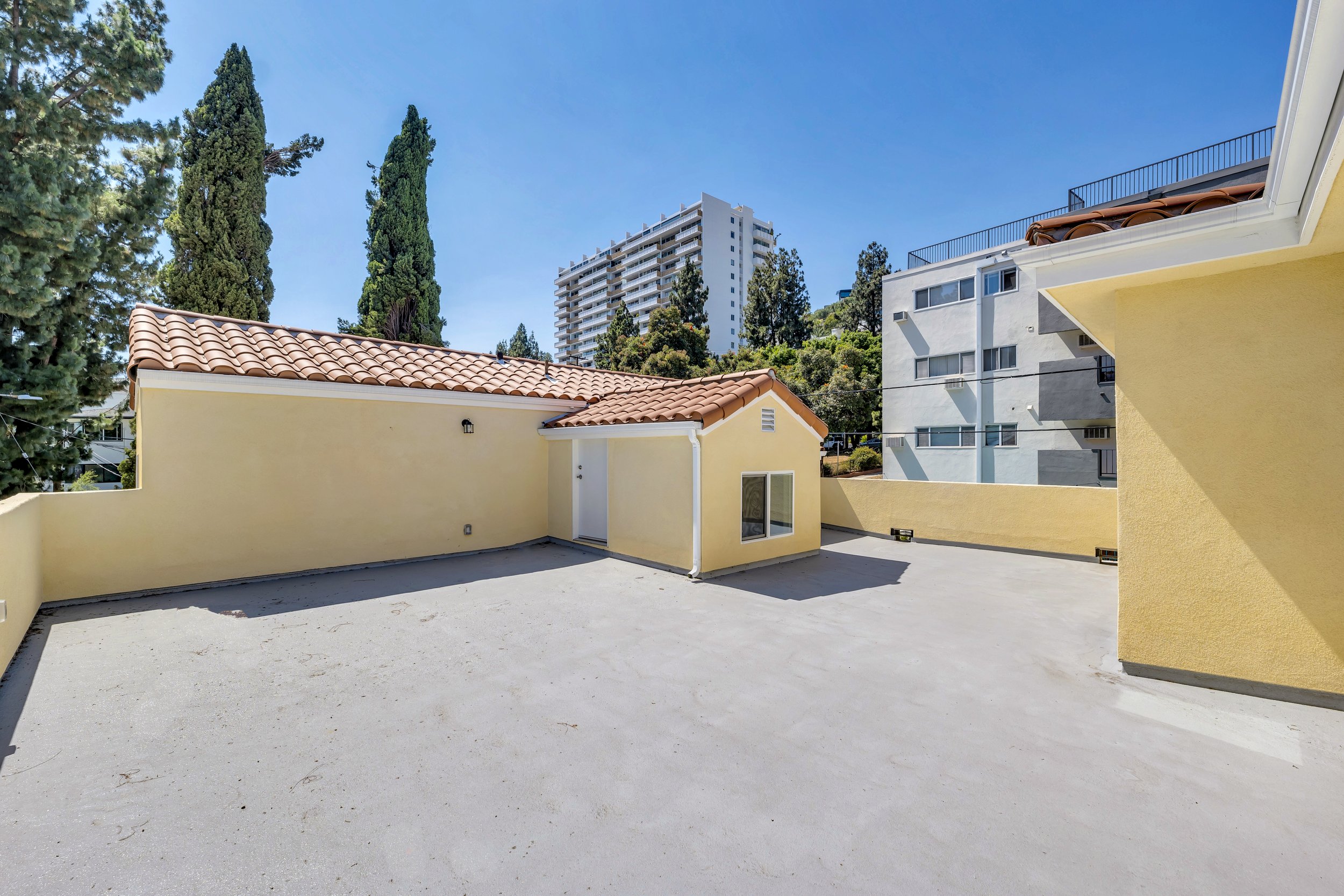 Empty outdoor rooftop patio with a smooth concrete floor, beige and yellow walls, and a view of tall trees and modern high-rise apartments in the background under a clear blue sky.