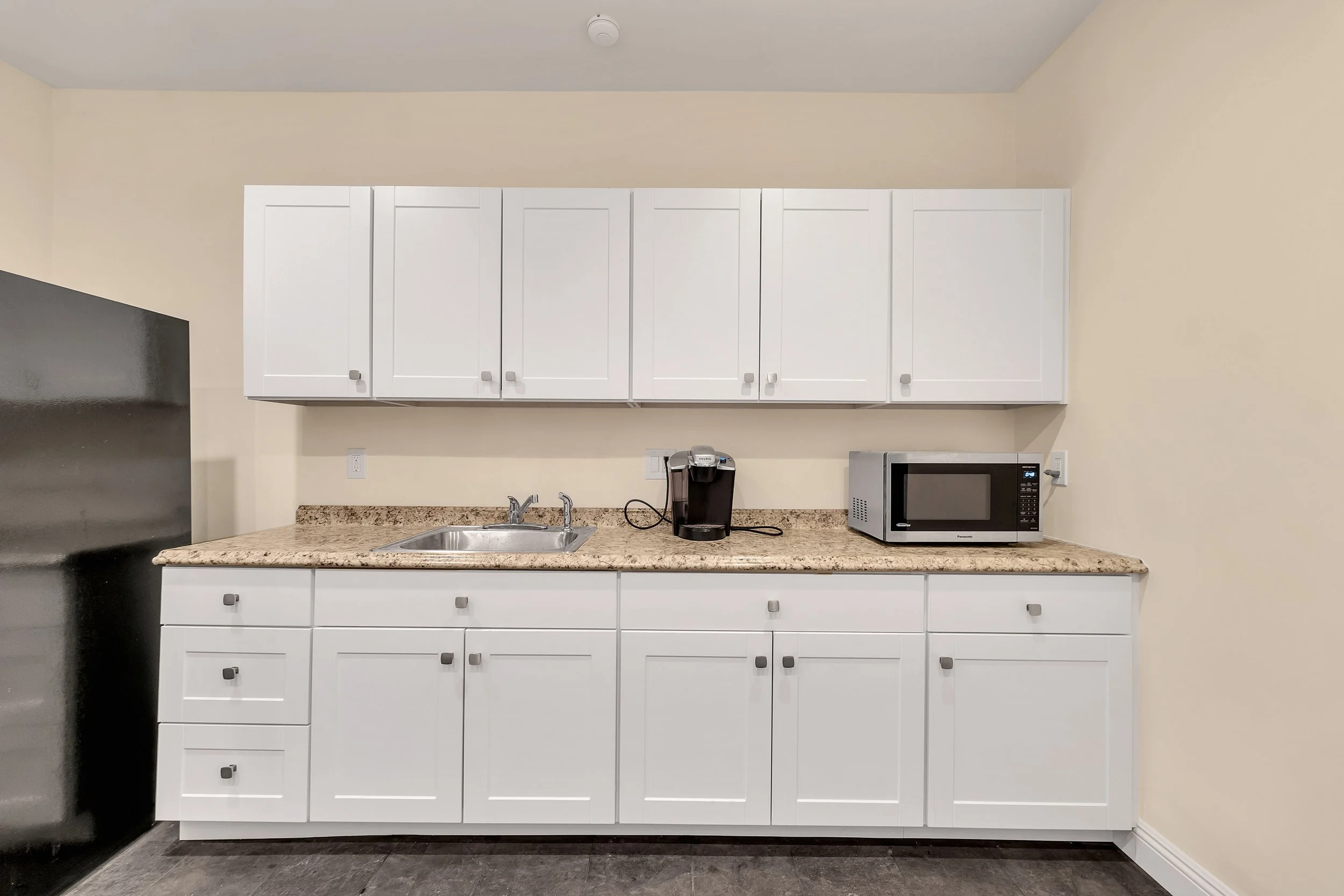 A kitchen counter with white cabinets above and below, a granite countertop, a stainless steel sink, a Keurig coffee maker, a microwave, and electrical outlets on the wall.