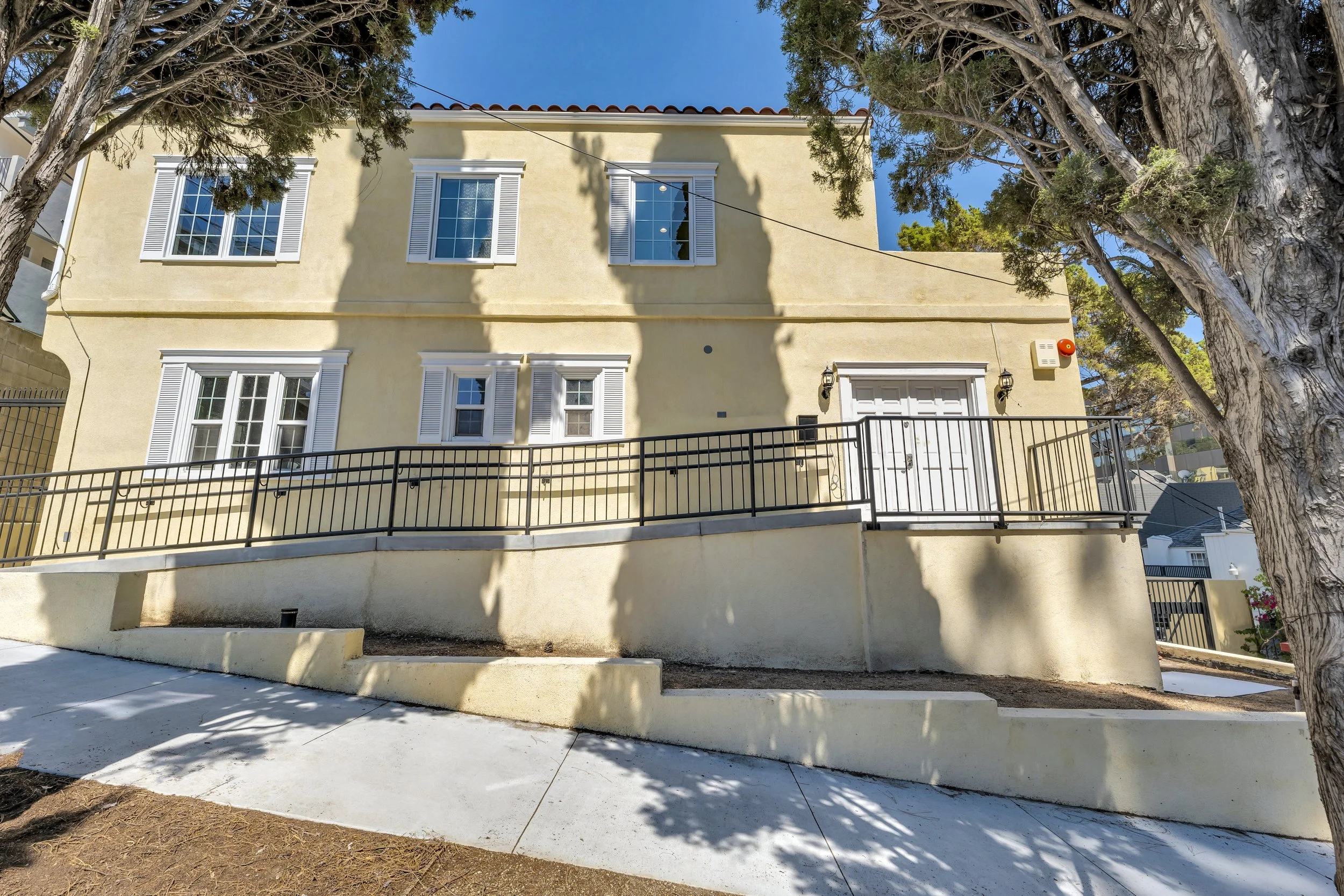 A two-story yellow house with white window shutters and a white front door, enclosed by a black metal fence. Large trees shade parts of the house, and the sky is blue.