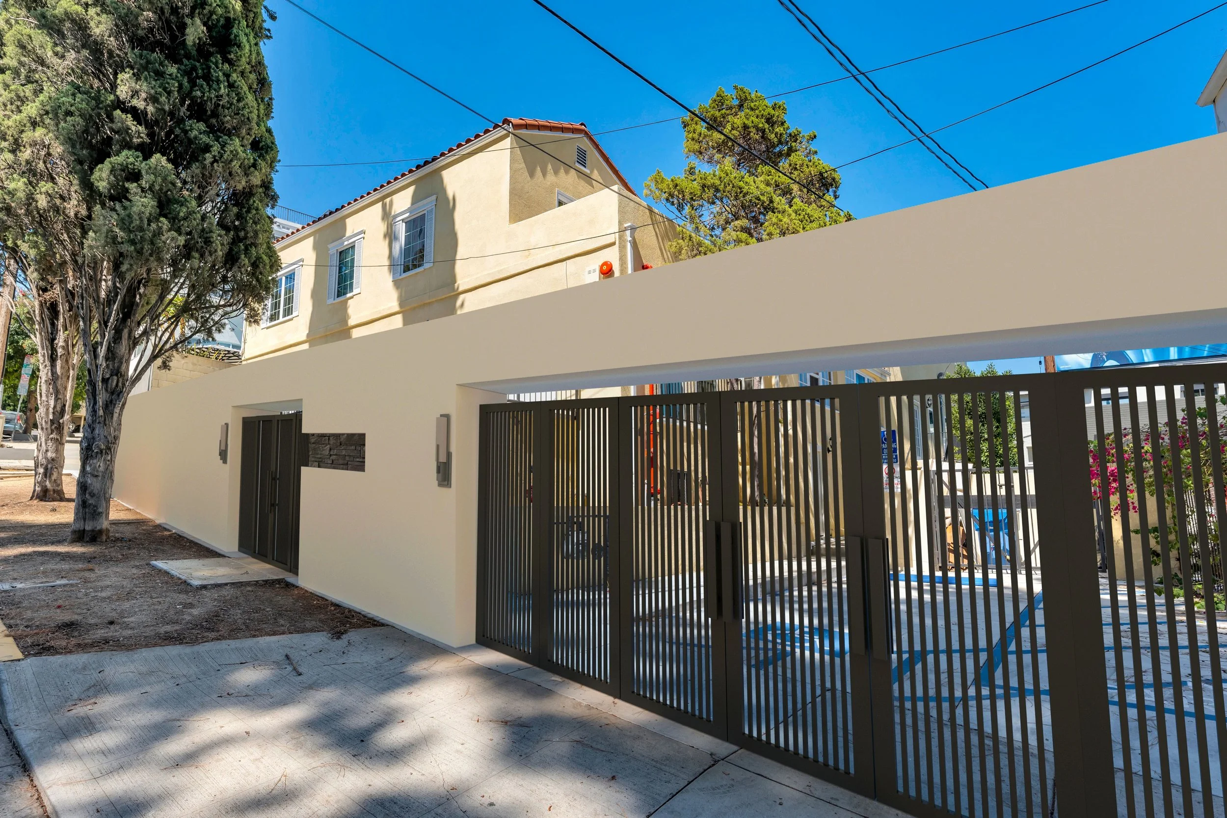 A modern white wall with a black vertical slat gate, adjacent to a sidewalk, with a yellow stucco house with a red tile roof and white framed windows behind, topped with a bright blue sky and green trees.