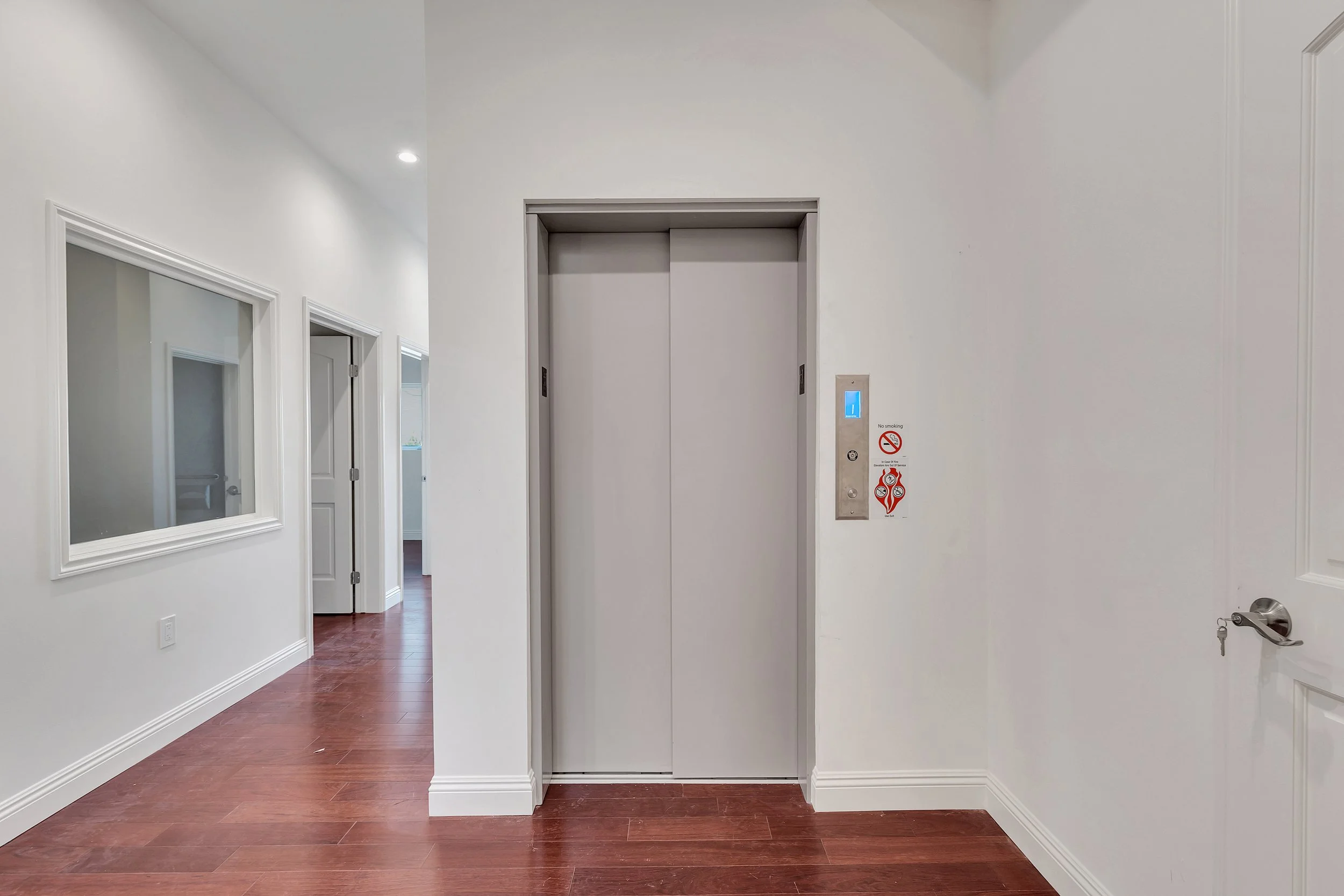 An elevator door with a digital floor indicator, and various signs including no smoking and fire safety signs, in a clean, white hallway with wooden flooring.