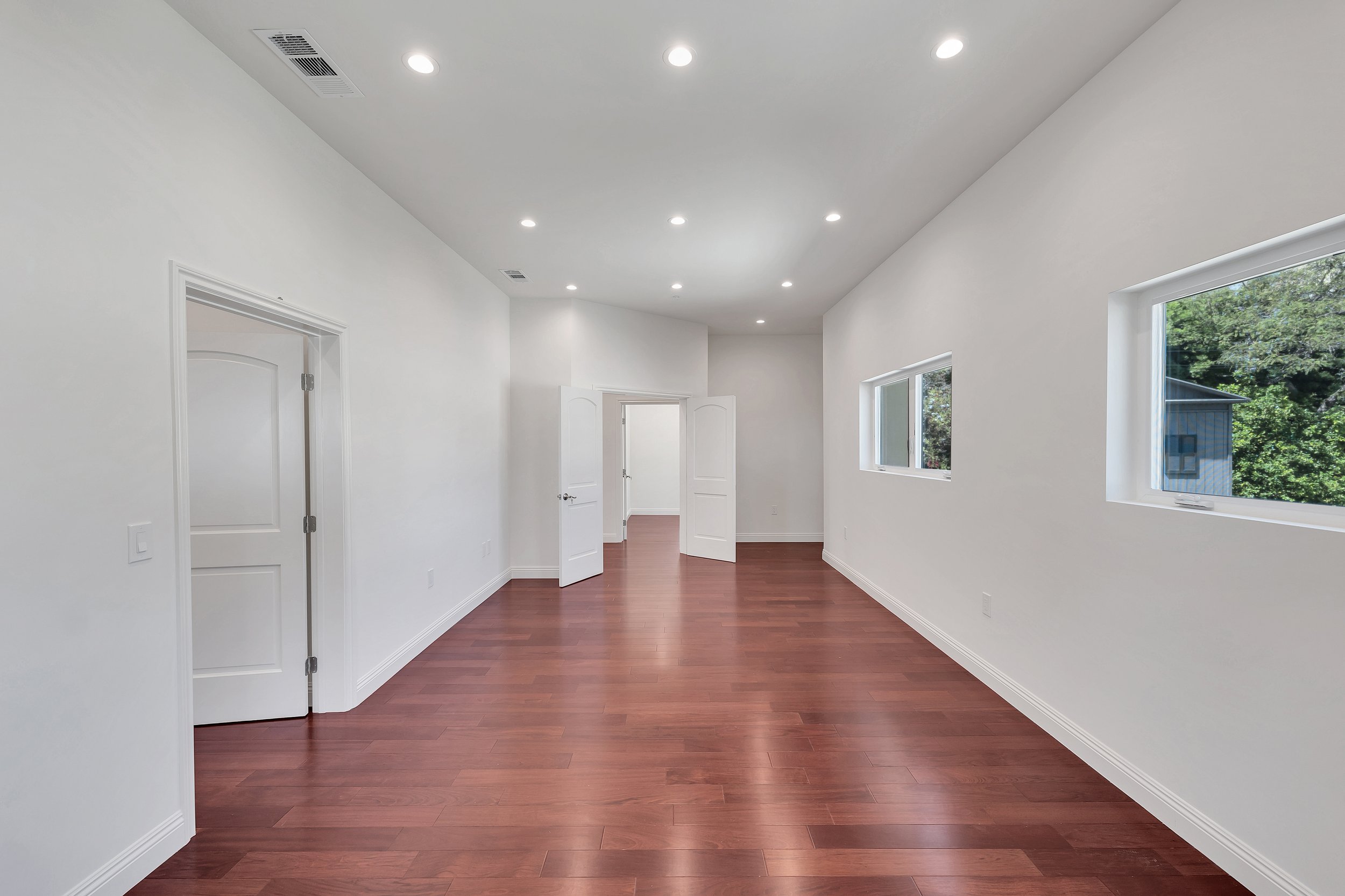 Empty room with white walls, hardwood floors, ceiling lights, and windows showing greenery outside.