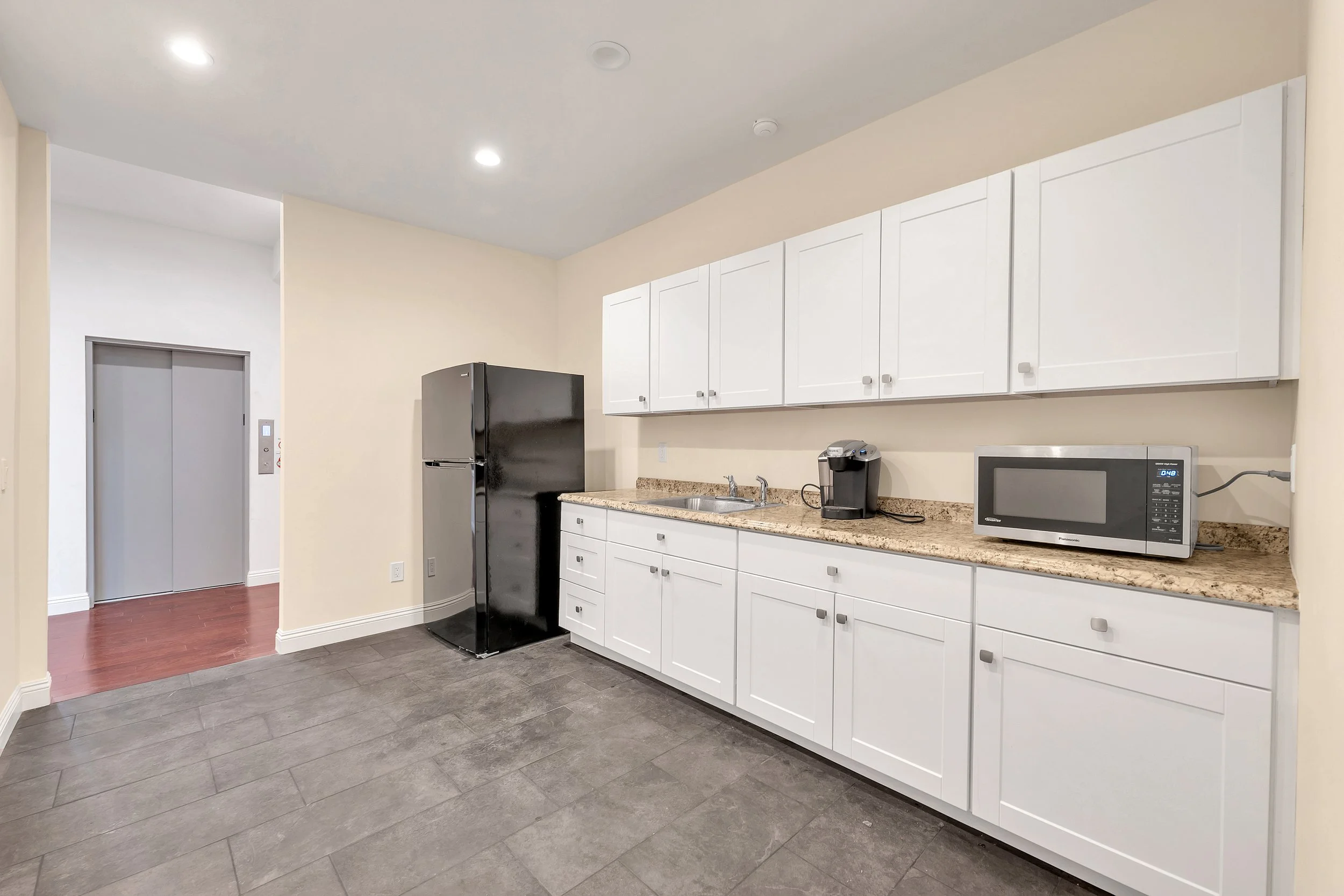 A kitchen with white cabinets, a granite countertop, a black refrigerator, a microwave, and a coffee maker. The floor is dark gray tile, and there are ceiling lights and a doorway to an elevator area.