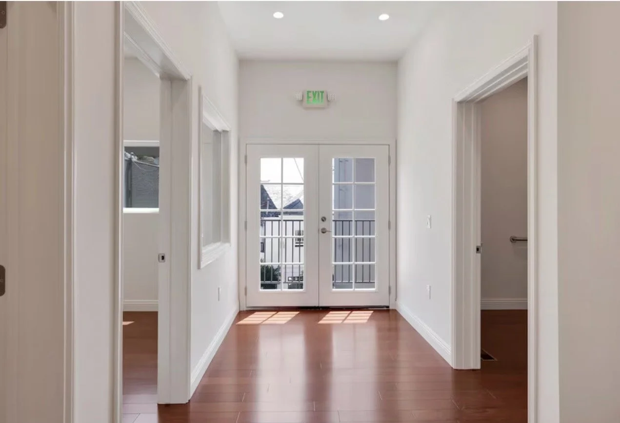 Entryway with glass-paneled double doors leading outside, white walls, and hardwood floors. There are doorways to rooms on each side and a balcony visible through the doors.