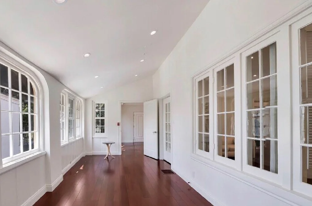Bright, empty room with white walls, multiple large windows with divided panes, hardwood floors, and a small table near the door.