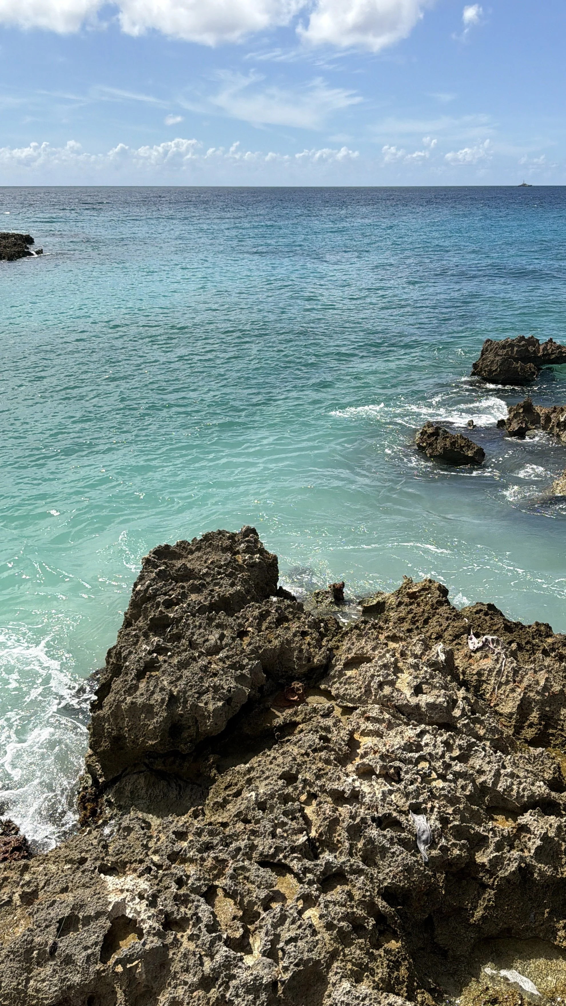 Bright blue ocean water with a rocky shoreline in the foreground under a partly cloudy sky.
