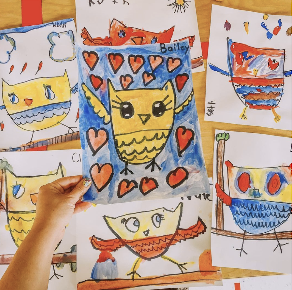 A student holding up a painted owl, with more student owl paintings pinned behind