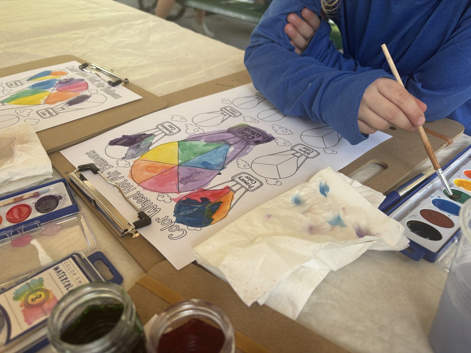 A child dipping a brush into a watercolor palette beside a color wheel worksheet