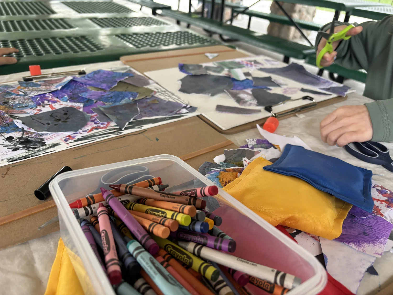 A tub of crayons in the foreground with children cutting and gluing paper collages behind