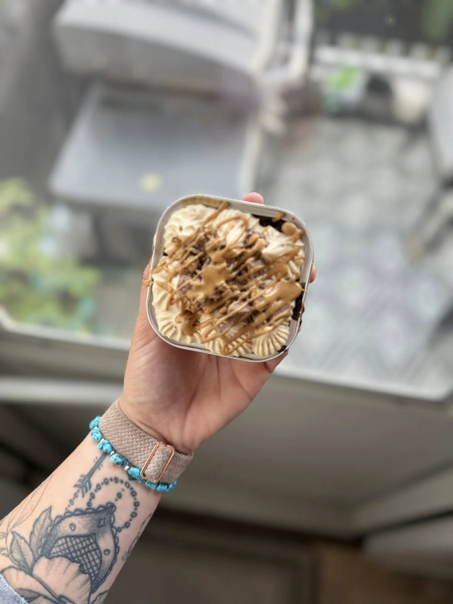 Person holding a tub of ice cream topped with caramel sauce and chocolate drizzle, with tattoos on their arm and a bracelet, standing indoors near a window.