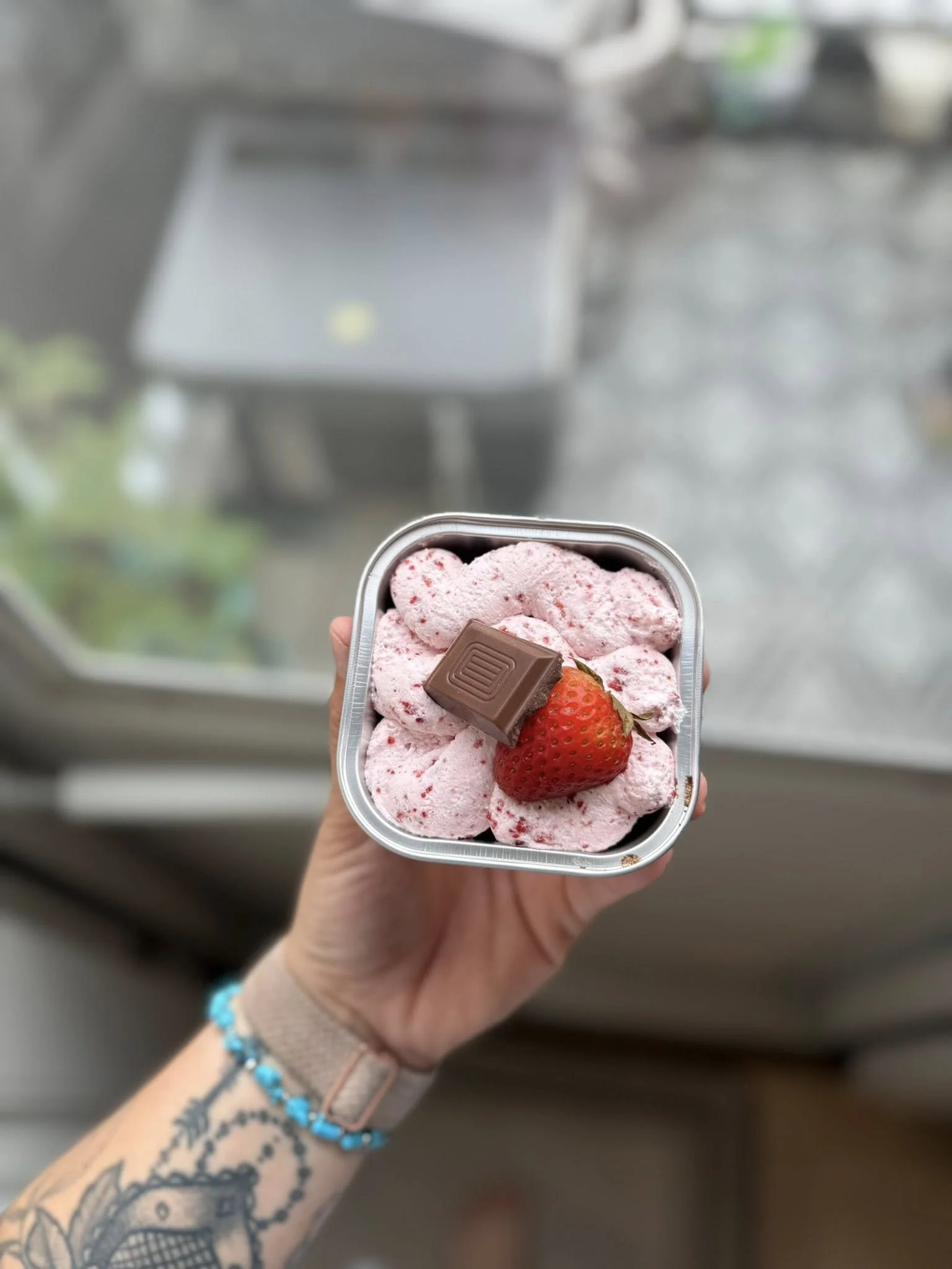 Hand holding a small square container of strawberry ice cream topped with a chocolate piece and a whole strawberry, with a tattooed arm and a blue bracelet, against a blurred background.