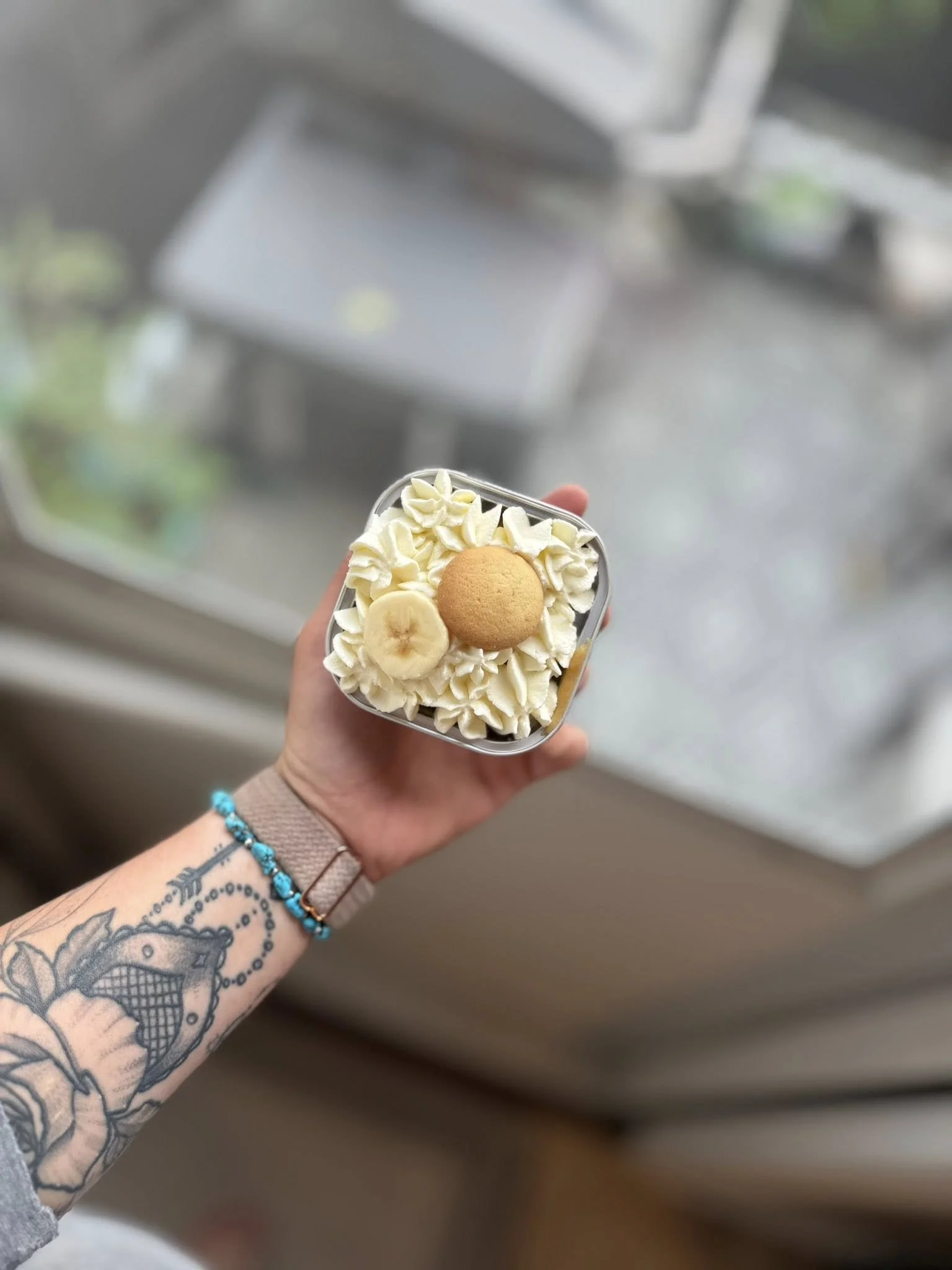 Hand holding a small square container of vanilla ice cream topped with whipped cream, banana slices, a cookie, and white chocolate shavings, with a balcony and city view in the background.