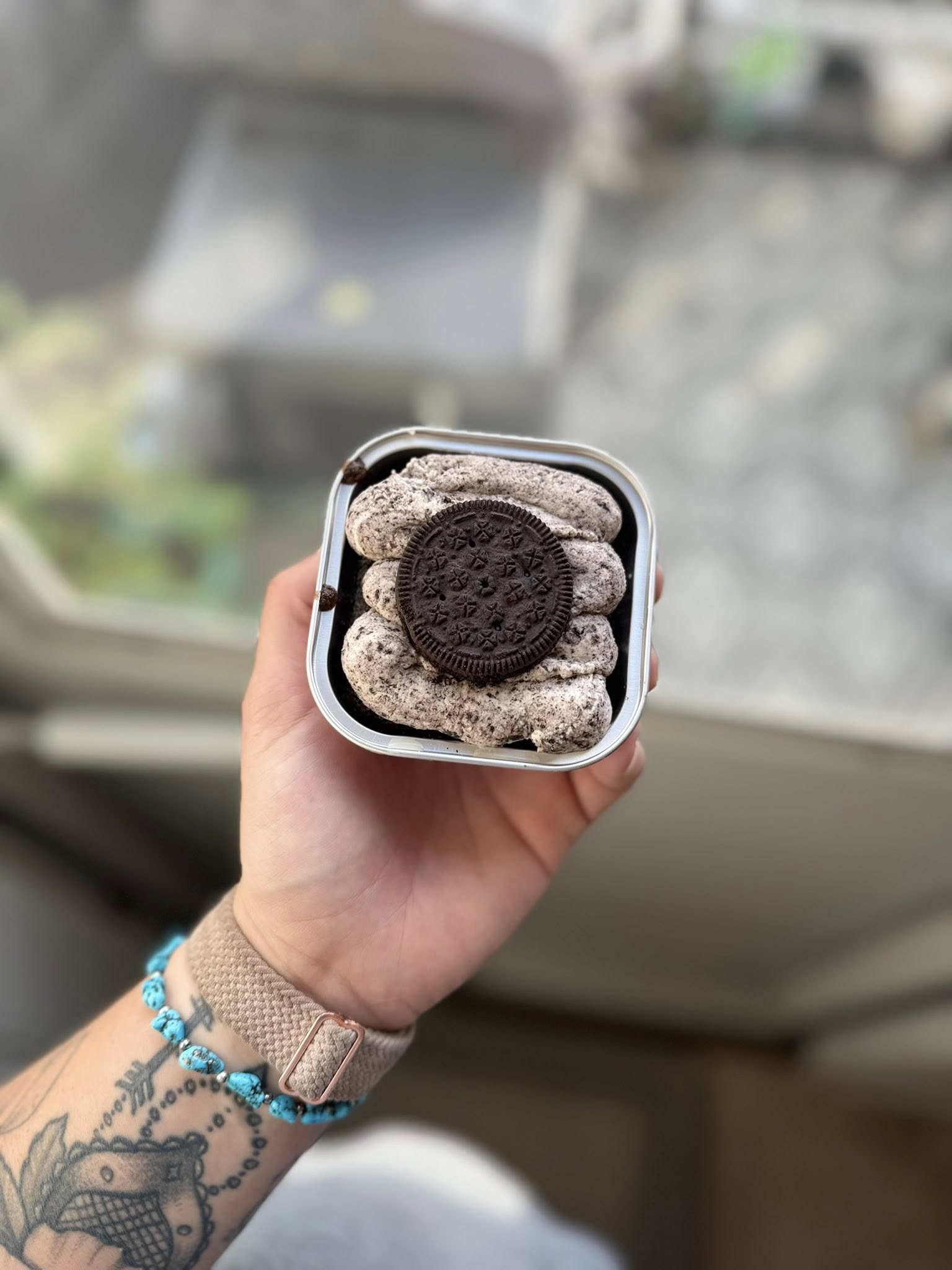 A hand holding a small container of cookies topped with an Oreo cookie. The person has a tattoo and is wearing a turquoise beaded bracelet and a beige wristband. The background is blurred and shows a window and some outdoor scenery.