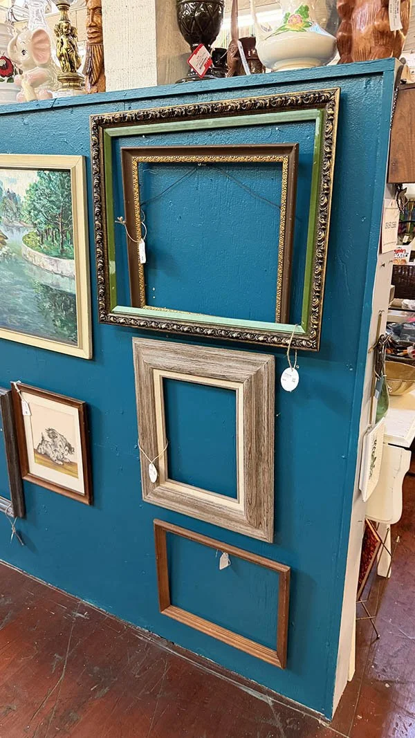 Empty antique frames hung on a teal wall alongside framed art at an antique dealer