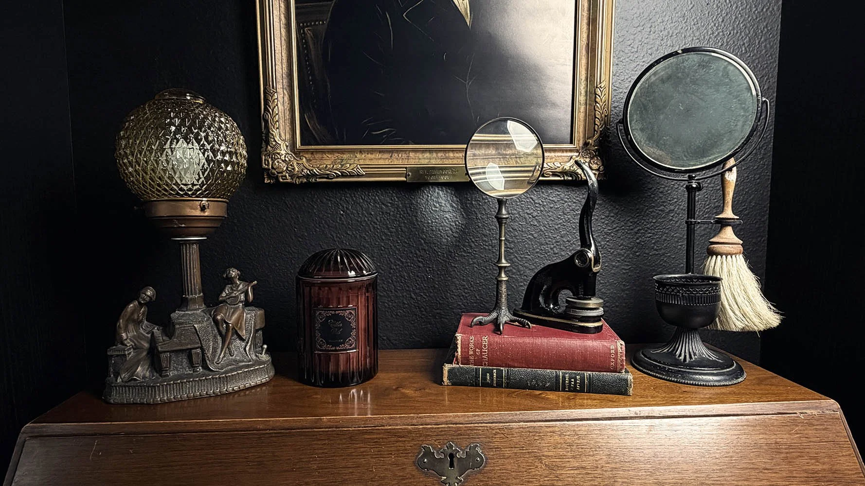 A collected antique desk vignette with a brass table lamp, glass candle jar, magnifying glass, antique desk seal press, and shaving mirror against a dark wall