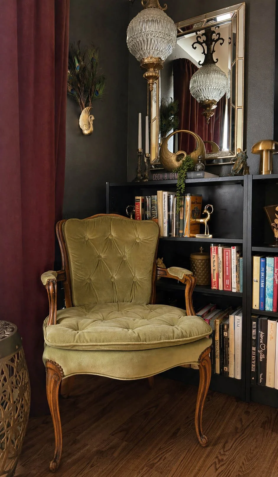 Green tufted armchair beside a black bookcase, with a glass pendant light and a gilt-framed mirror in a dark study.