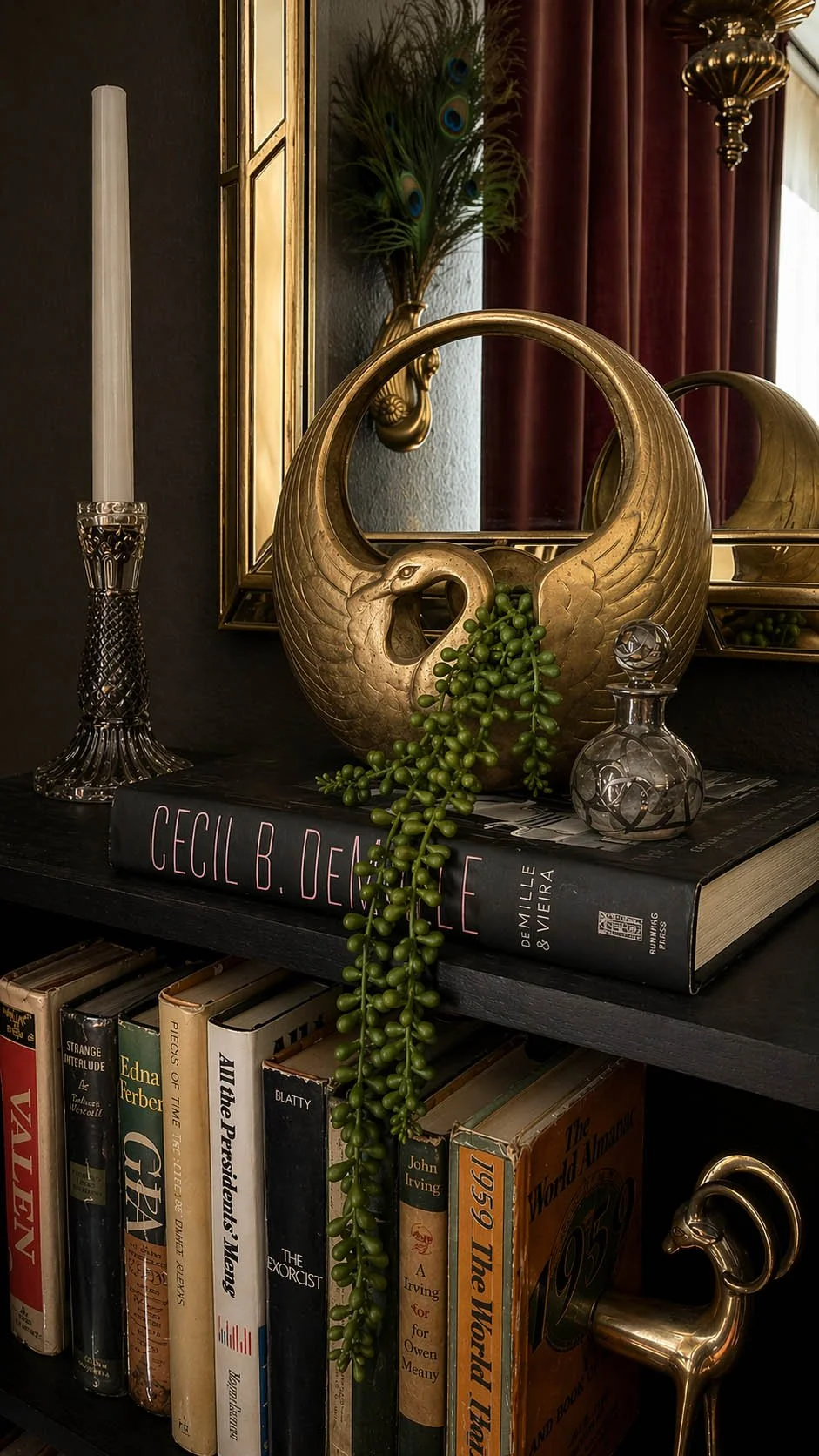 Brass swan basket with trailing string of pearls plant on a black bookshelf beside a Cecil B. DeMille coffee table book and crystal candlestick