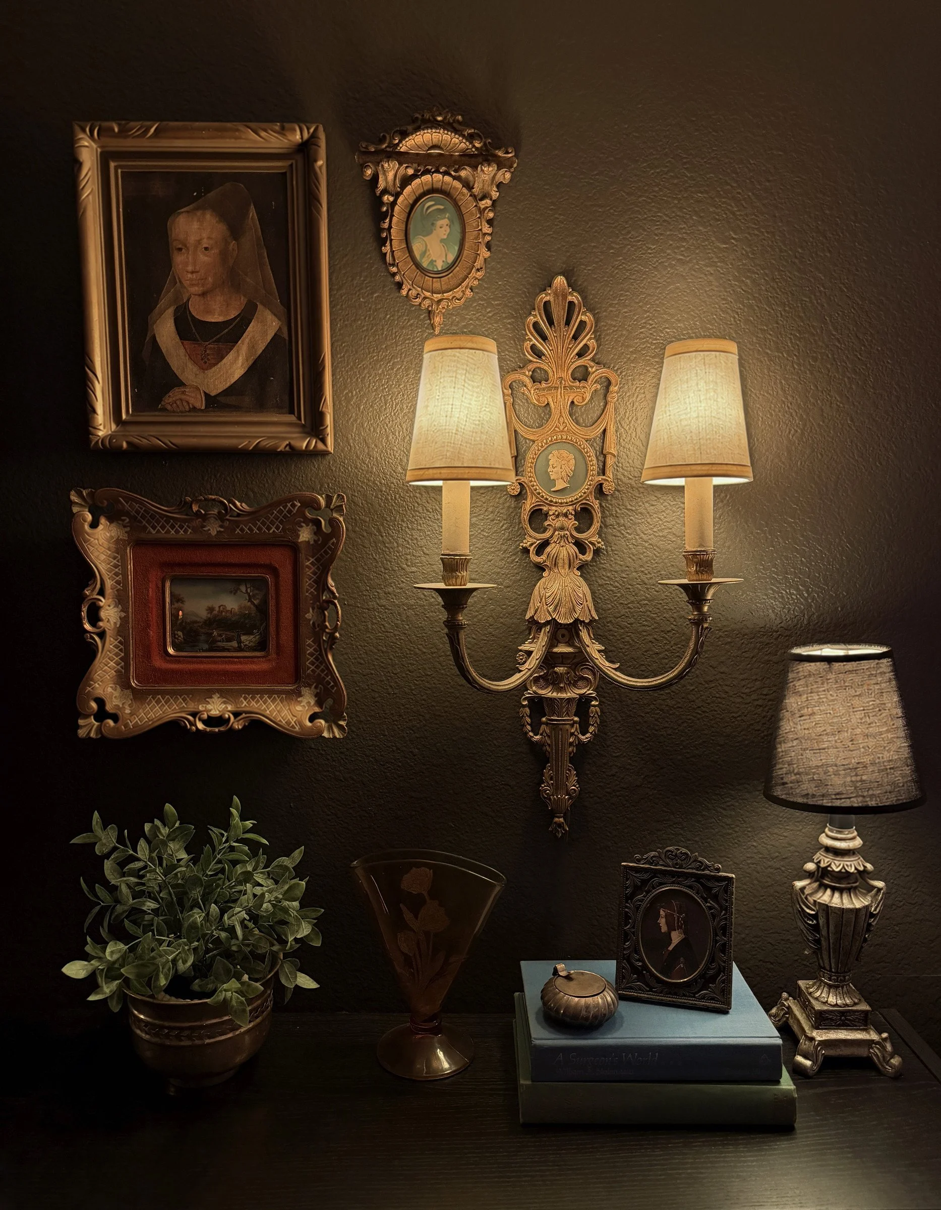 Brass wall sconce with two lit shades beside gilt-framed portraits above a dark shelf with a small lamp.