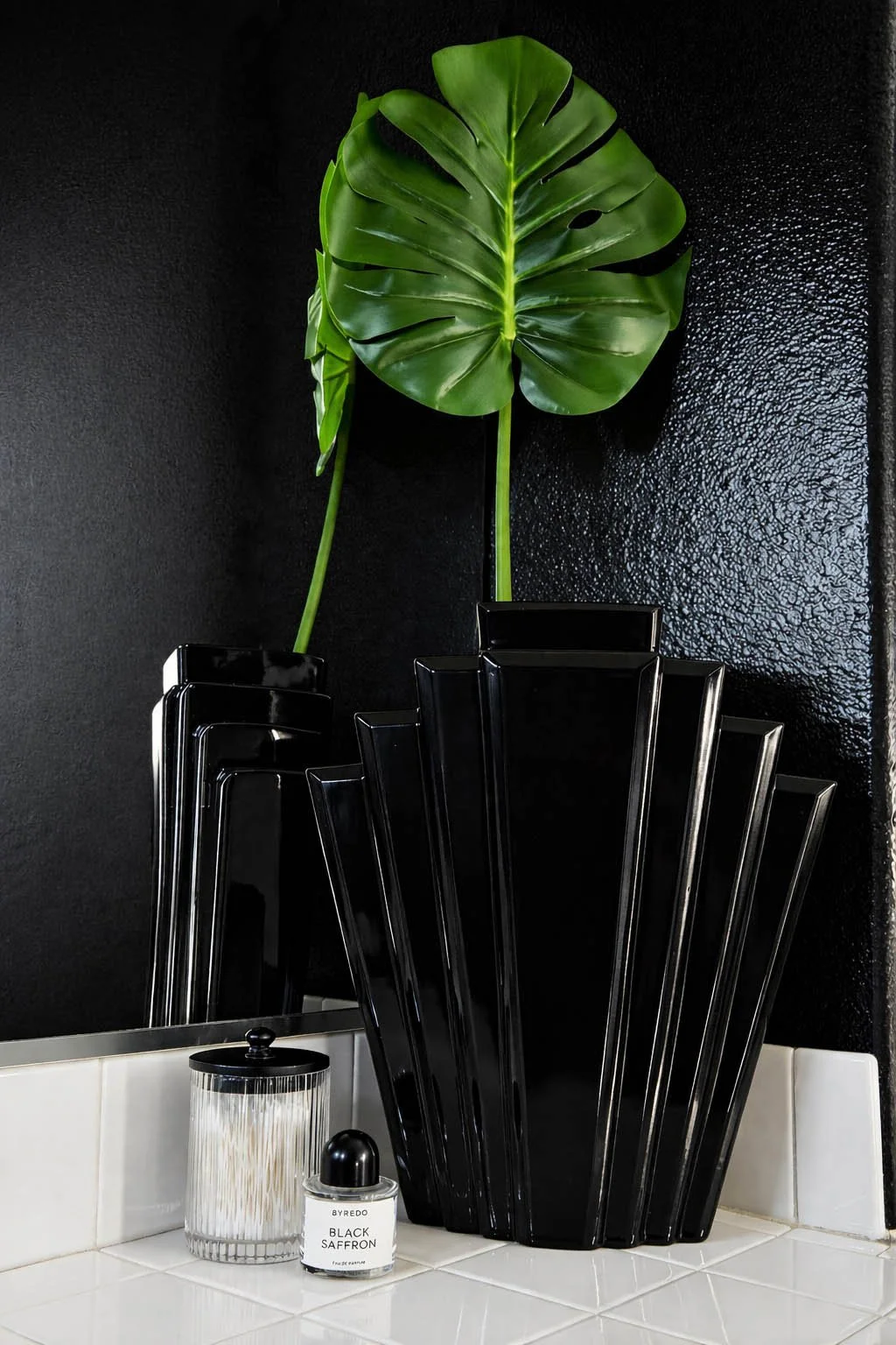 Pair of glossy black Art Deco fan vases with a single monstera leaf on white tile beside a Byredo Black Saffron perfume bottle