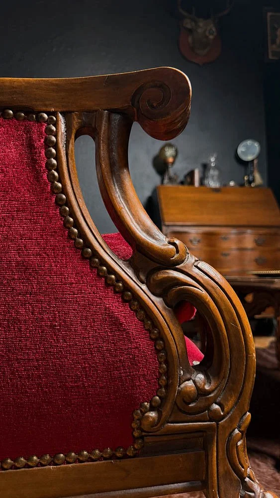 Detail of a carved wood wingback chair's scrollwork arm and red velvet upholstery, with an antique secretary desk in the background.