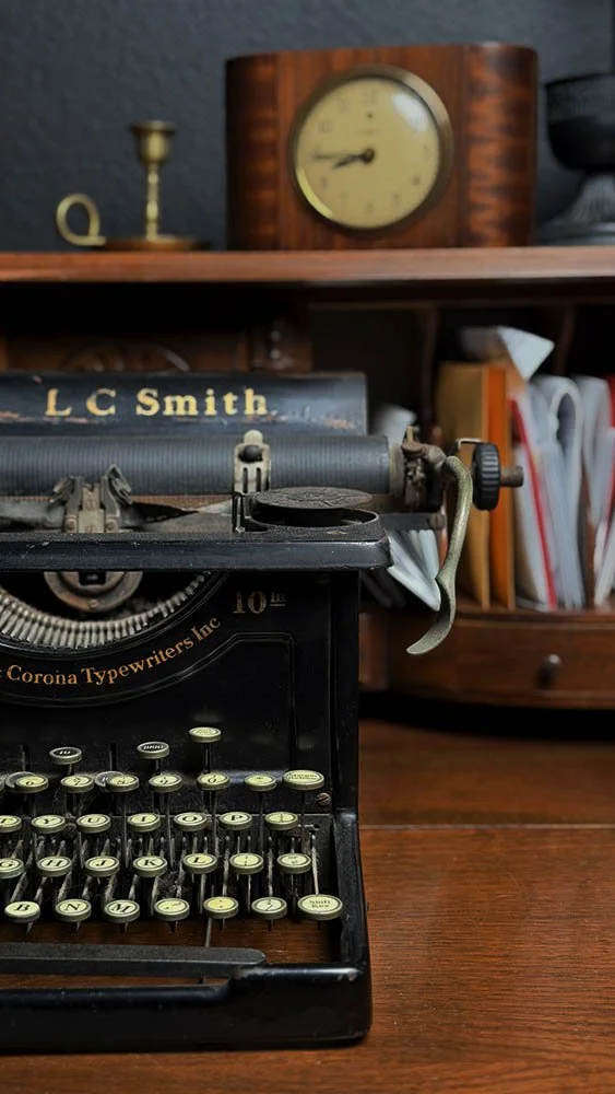 Vintage L.C. Smith typewriter centered on an antique secretary desk in a dark academia home office, with a wood-cased clock and brass candlestick in the background.