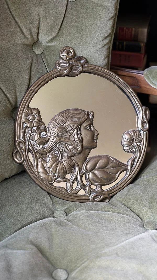 Round Art Nouveau brass mirror with a carved maiden profile against a sage green tufted chair