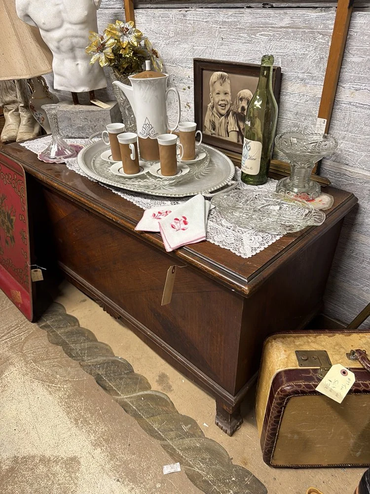 Scratched 1934 Lane cedar chest in an antique store, covered with glassware, a coffee set, and lace