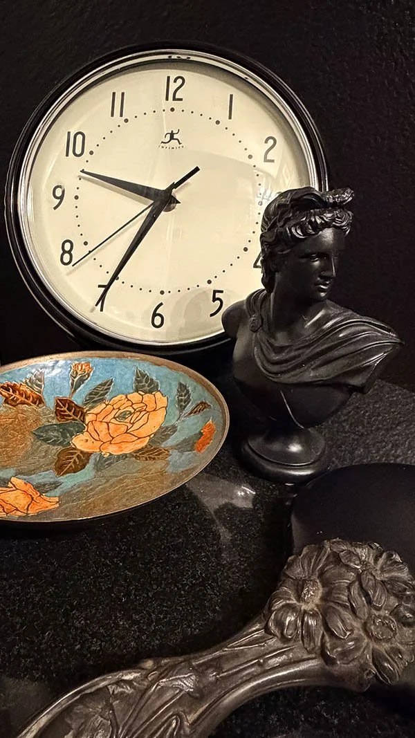 A round wall clock with a black bezel and cream face set against a black bust, a hand-painted floral dish, and an ornate silver frame, styled for a black lacquer guest bathroom vignette