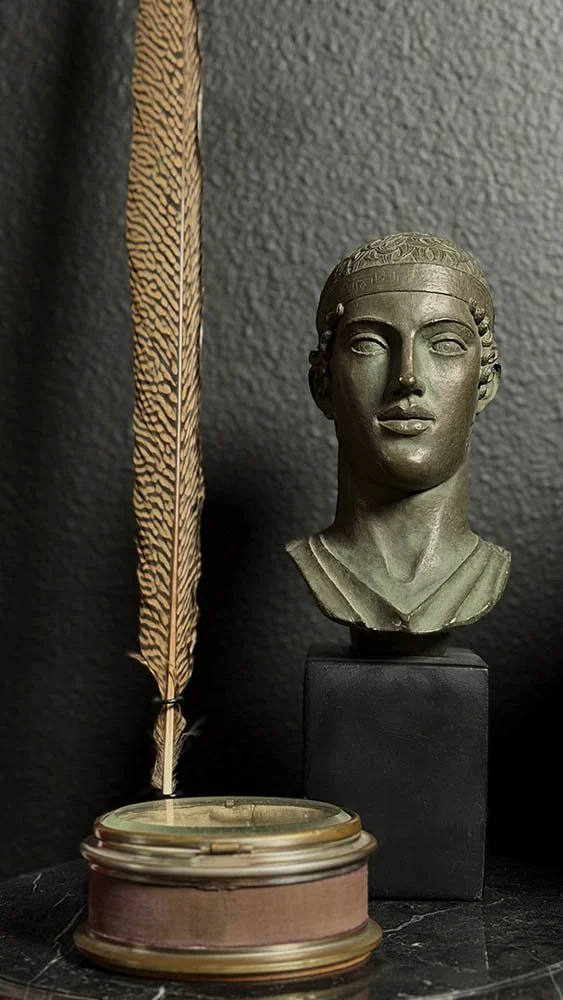 A bronze bust on a black pedestal beside a feather quill resting near a brass container.