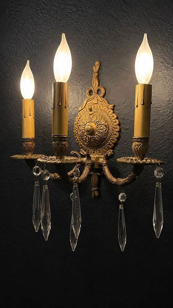 Three-arm brass wall sconce with hanging crystal drops, lit warmly against a dark textured wall.