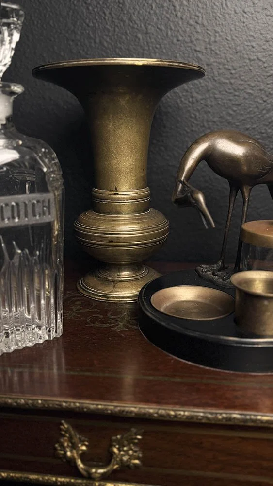 Brass trumpet vase, etched crystal Scotch decanter, and brass wading bird figurine on a dark wood desk
