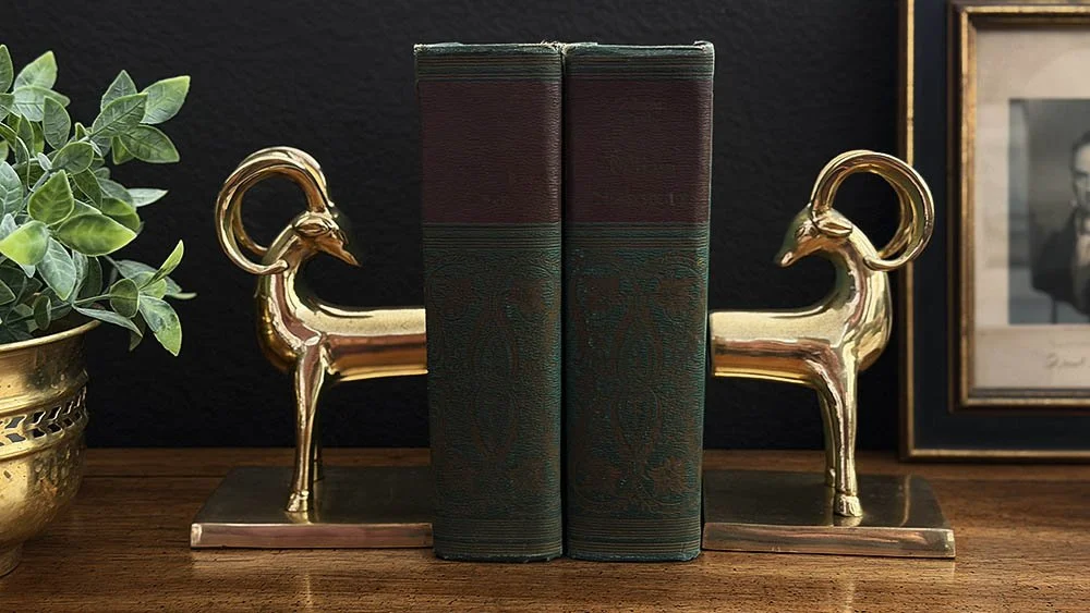 Pair of brass ibex bookends flanking antique leather bound books on a dark study shelf
