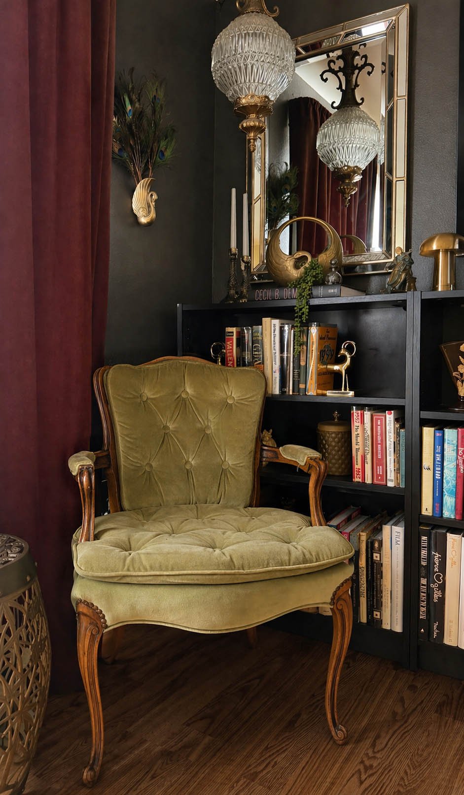 Sage velvet tufted armchair with carved walnut frame beside a dark bookshelf, crystal globe chandelier and peacock feather wall sconce in a moody eclectic interior