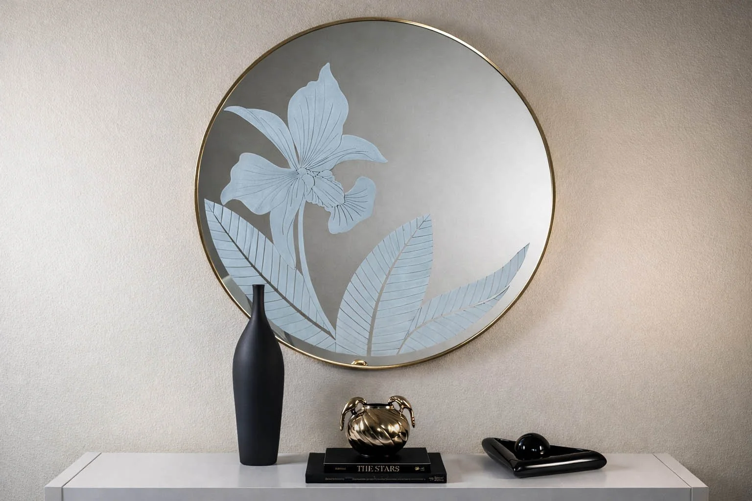 Large round gold-framed mirror with etched flower motif above a white console table styled with a matte black vase, gold ram vessel and art books