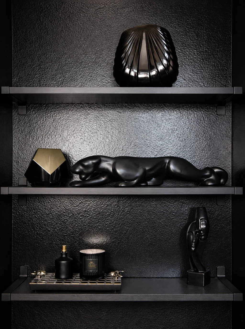 Three black shelves against a textured black wall styled with a matte black shell sculpture, prowling panther figurine, brass geometric object, luxury candle on a brass tray and abstract face sculpture