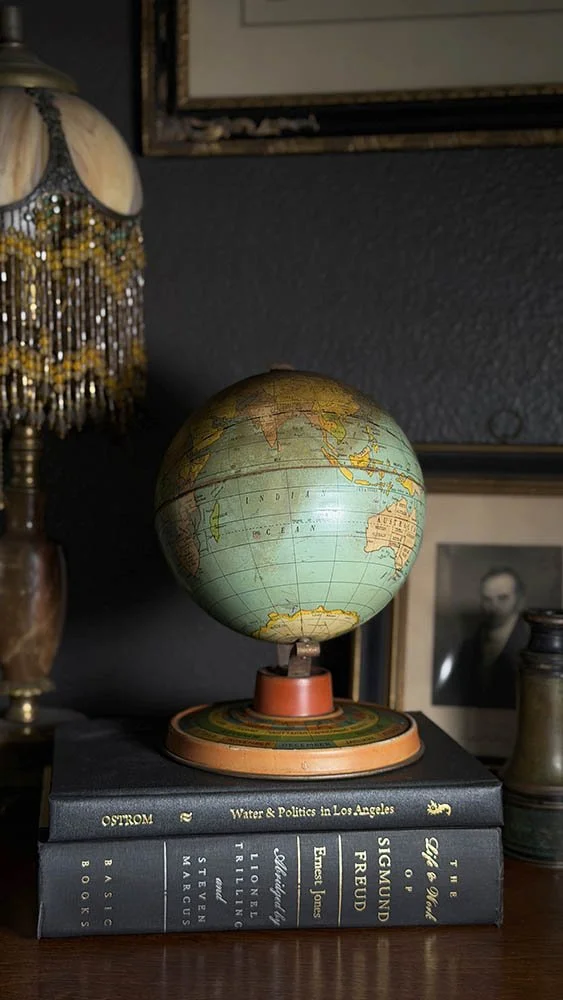Antique globe styled on a stack of books beside a beaded lamp and vintage portrait in a dark academia study corner