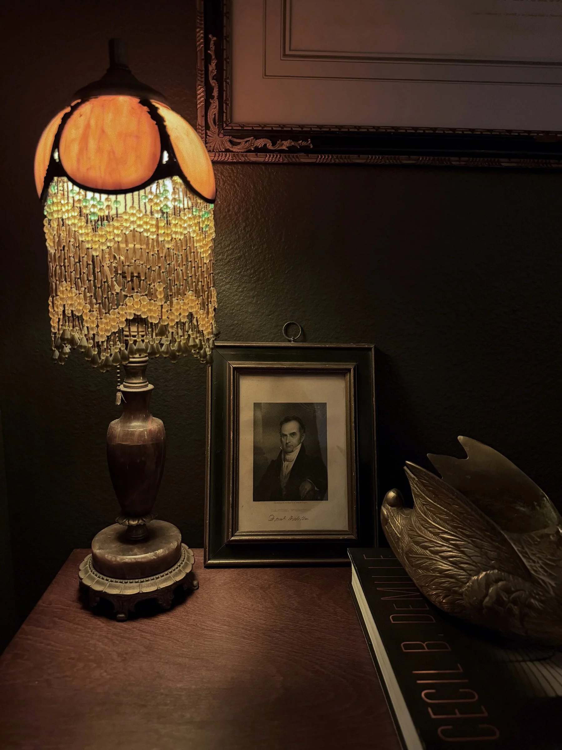 Beaded fringe table lamp lit beside a framed portrait print on a dark table, with a brass bird bowl at the edge of the frame.