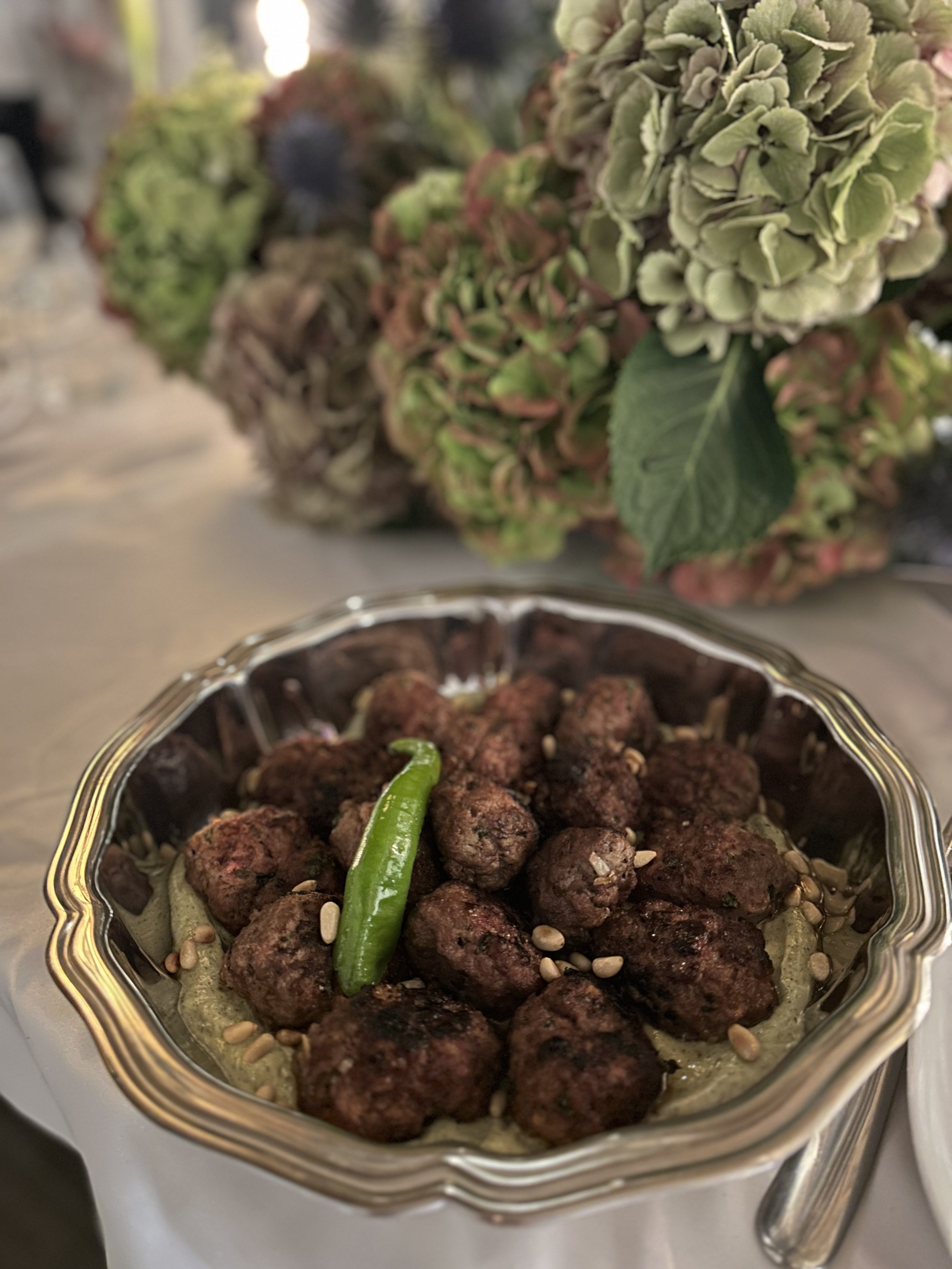 Un plat de boulettes de viande avec une chili vert en garniture, dans un plat en verre argenté, avec un bouquet de fleurs séchées en arrière-plan.