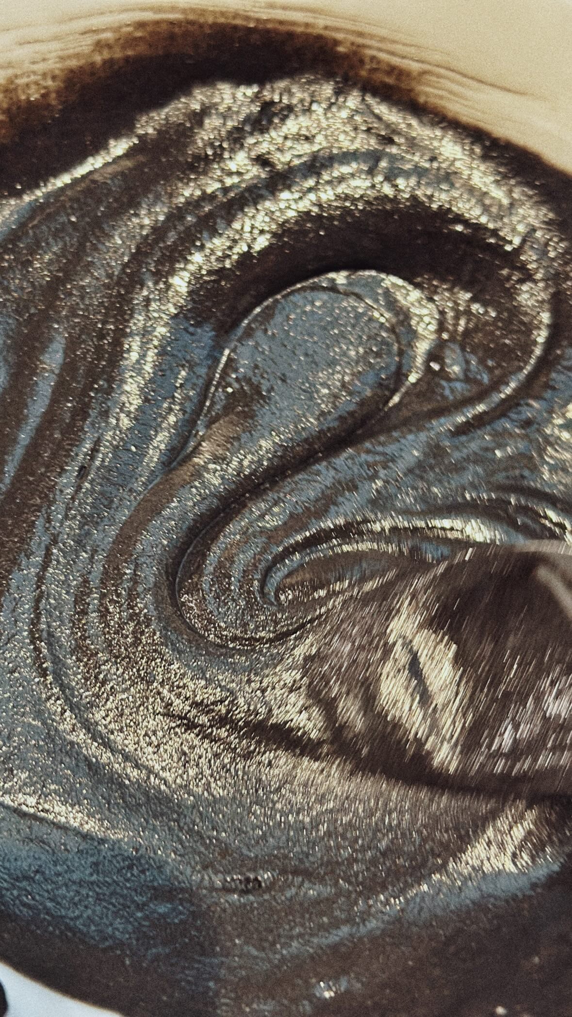 Une surface métallique noire, probablement une cuve ou un récipient, avec un liquide noir brillant ou une substance noire qui semble être en train de s'étaler ou de couler en spirale.
