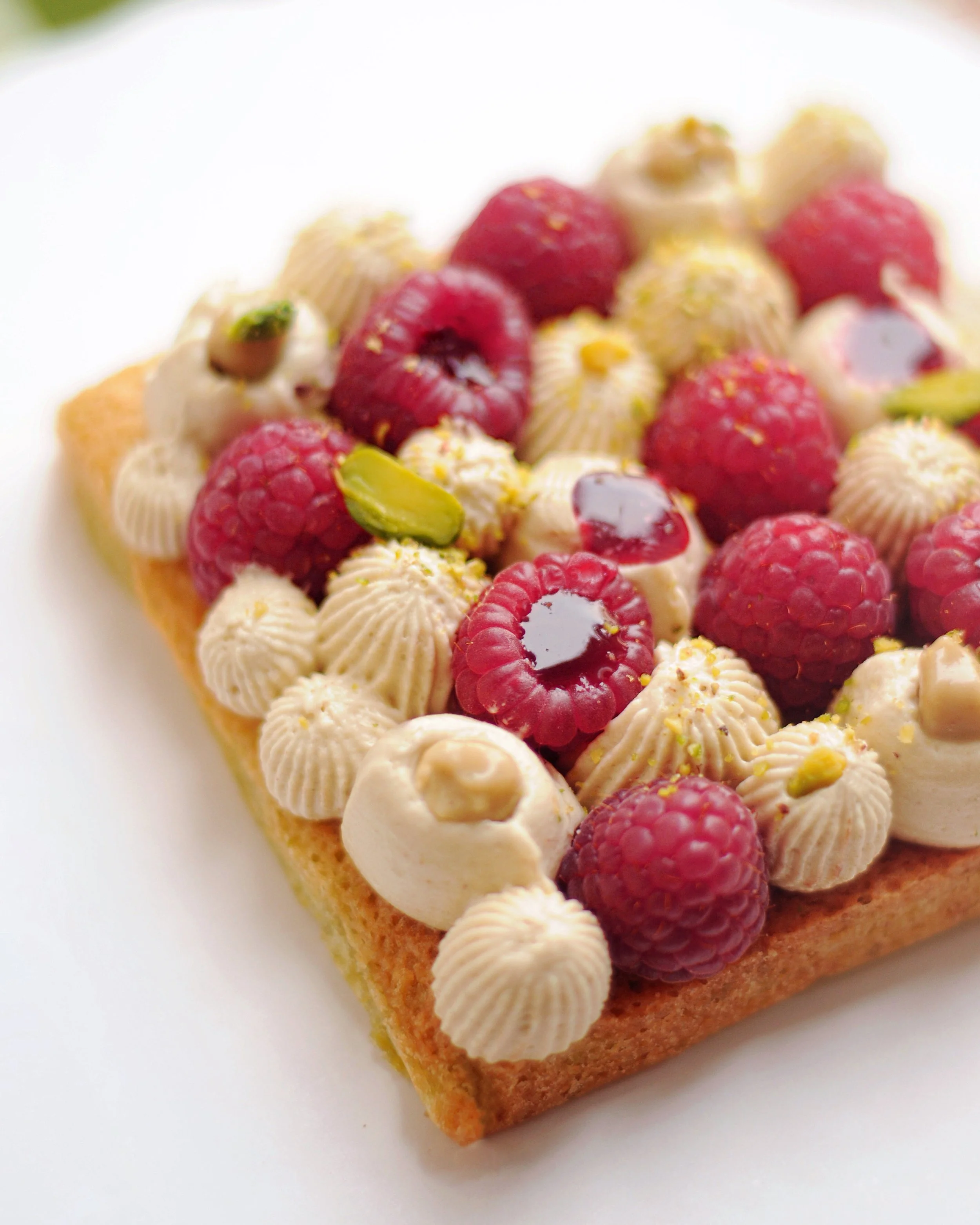 Financier pistache avec des framboises, des couches de crème, et des pistaches sur une base génoise, vu de côté.