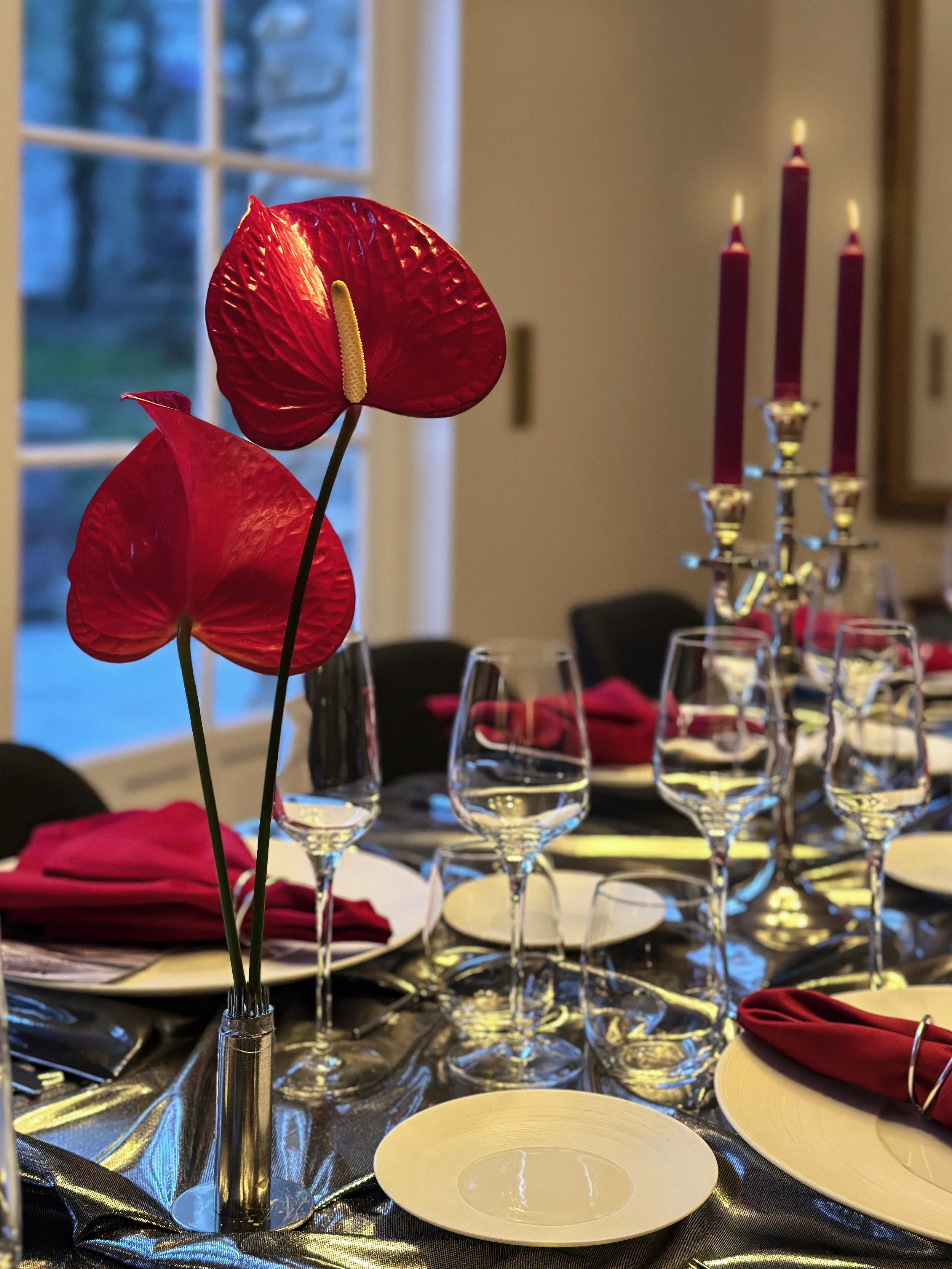 Une décoration de table avec un flambeau et des fleurs artificielles rouges, des verres à vin, des assiettes blanches et des serviettes rouges pliées. En arrière-plan, un chandelier avec trois bougies rouges allumées, dans une salle avec fenêtre.