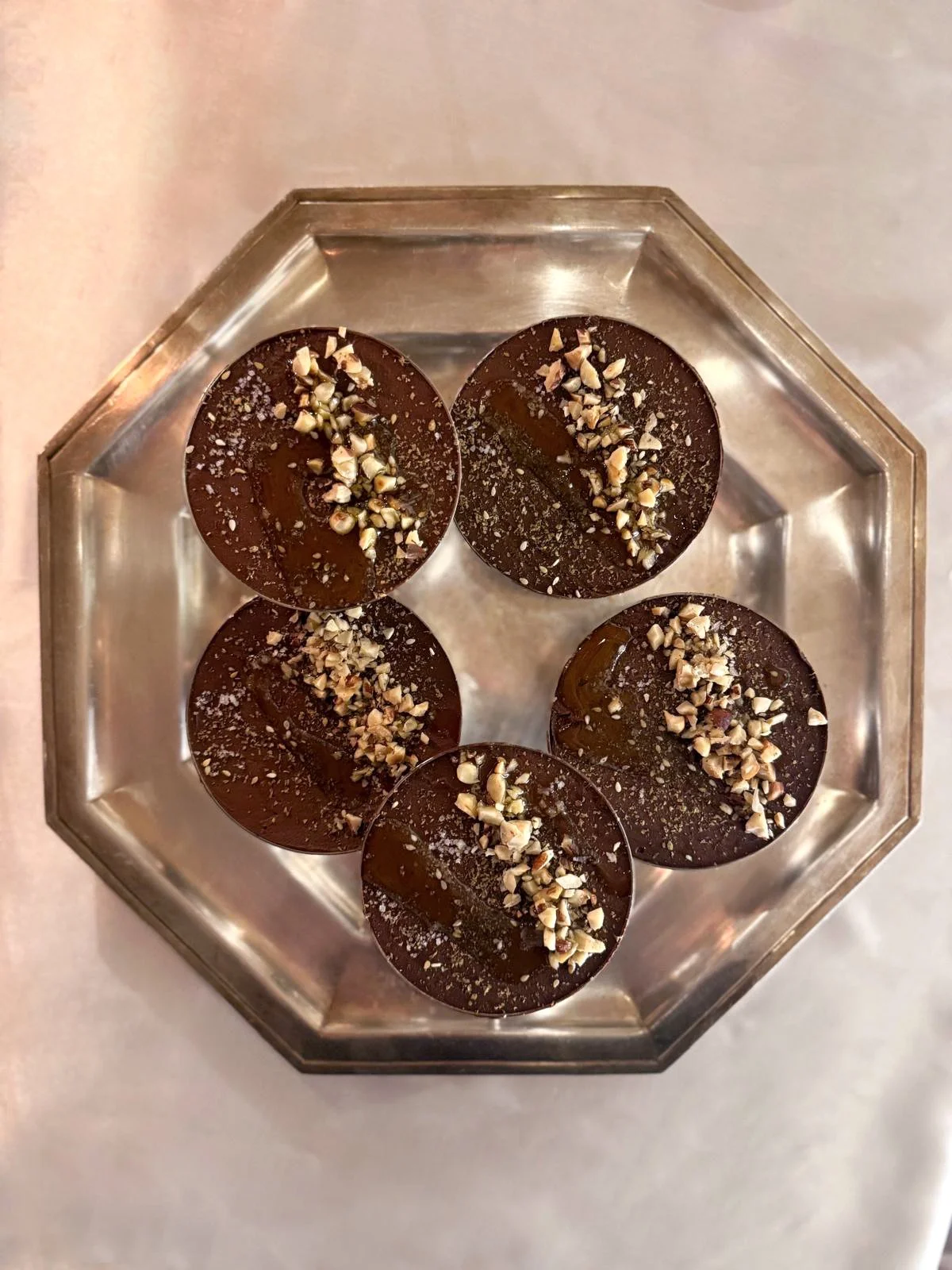 Cinq mousses au chocolat dans des coupes en inox avec des éclats de noix, huile d'olive, zaatar et fleur de sel sur le dessus, disposés dans un plat en métal.