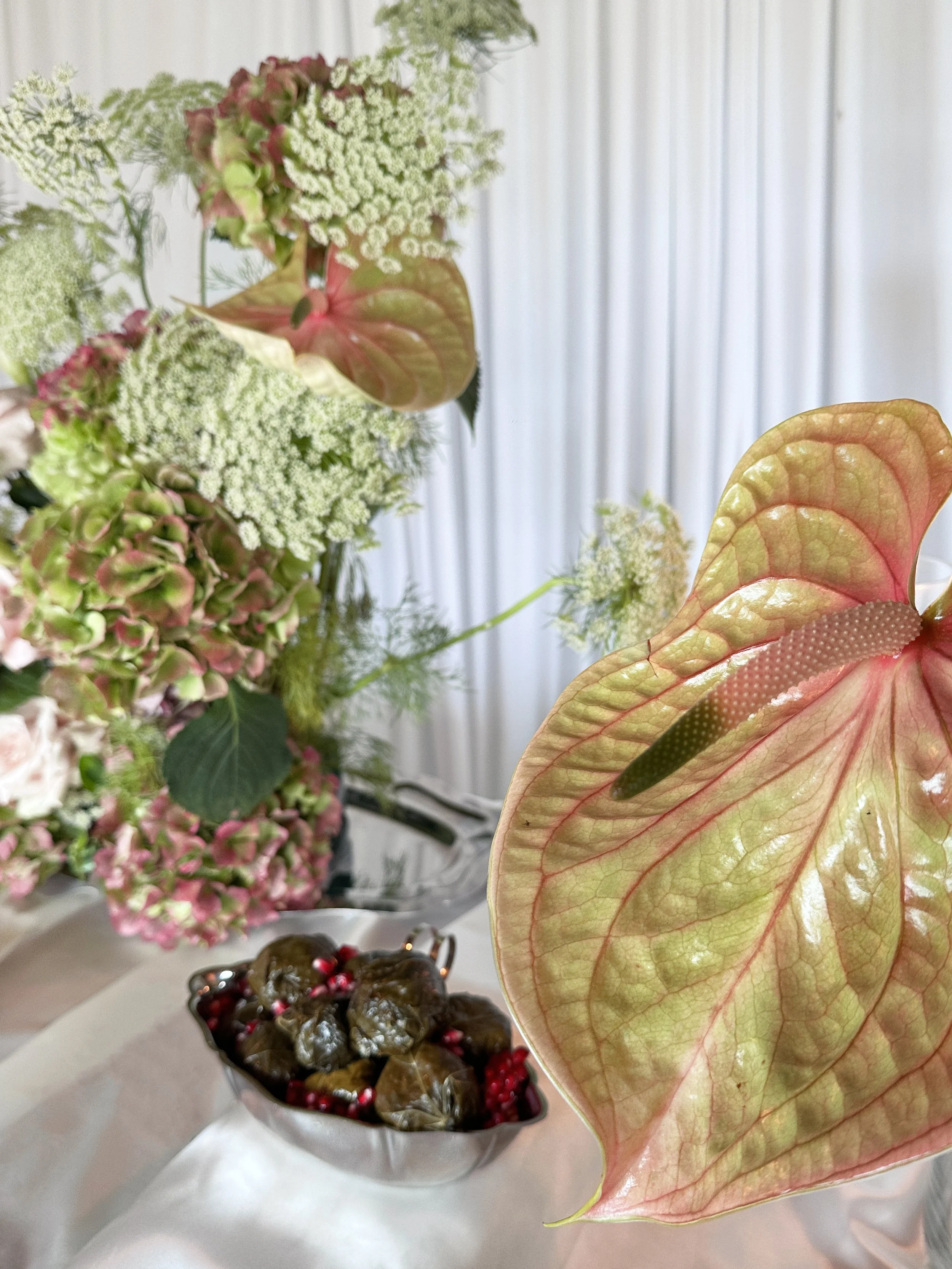 Un arrangement floral composé de fougères, d'hortensias et de caladiums, avec un bol contenant des boules de feuille de vigne et un bol avec des fruits rouges en arrière-plan.