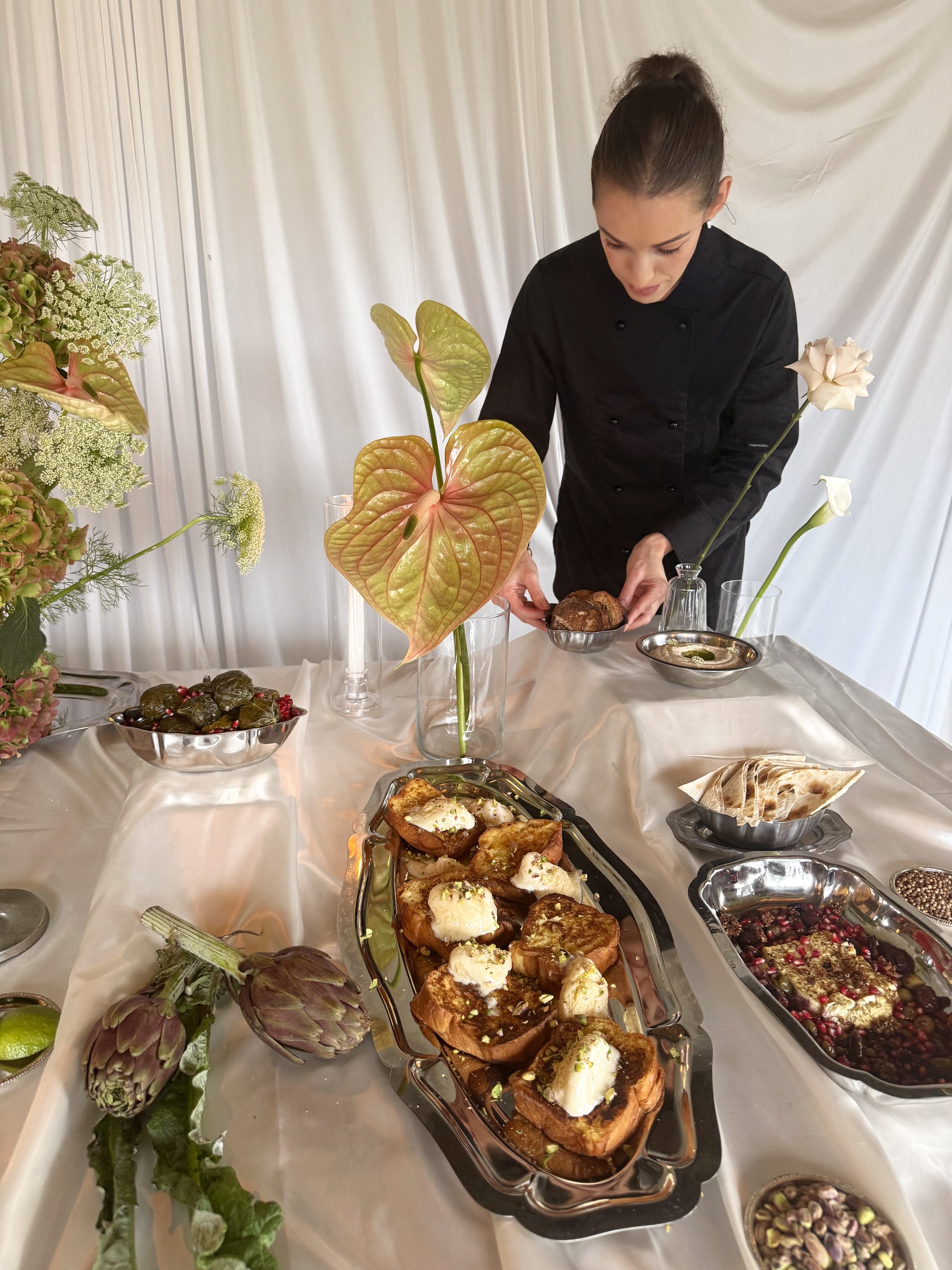 Une femme en uniforme de cuisine présente un plateau de nourriture, entourée de fleurs et de plats variés sur une table.
