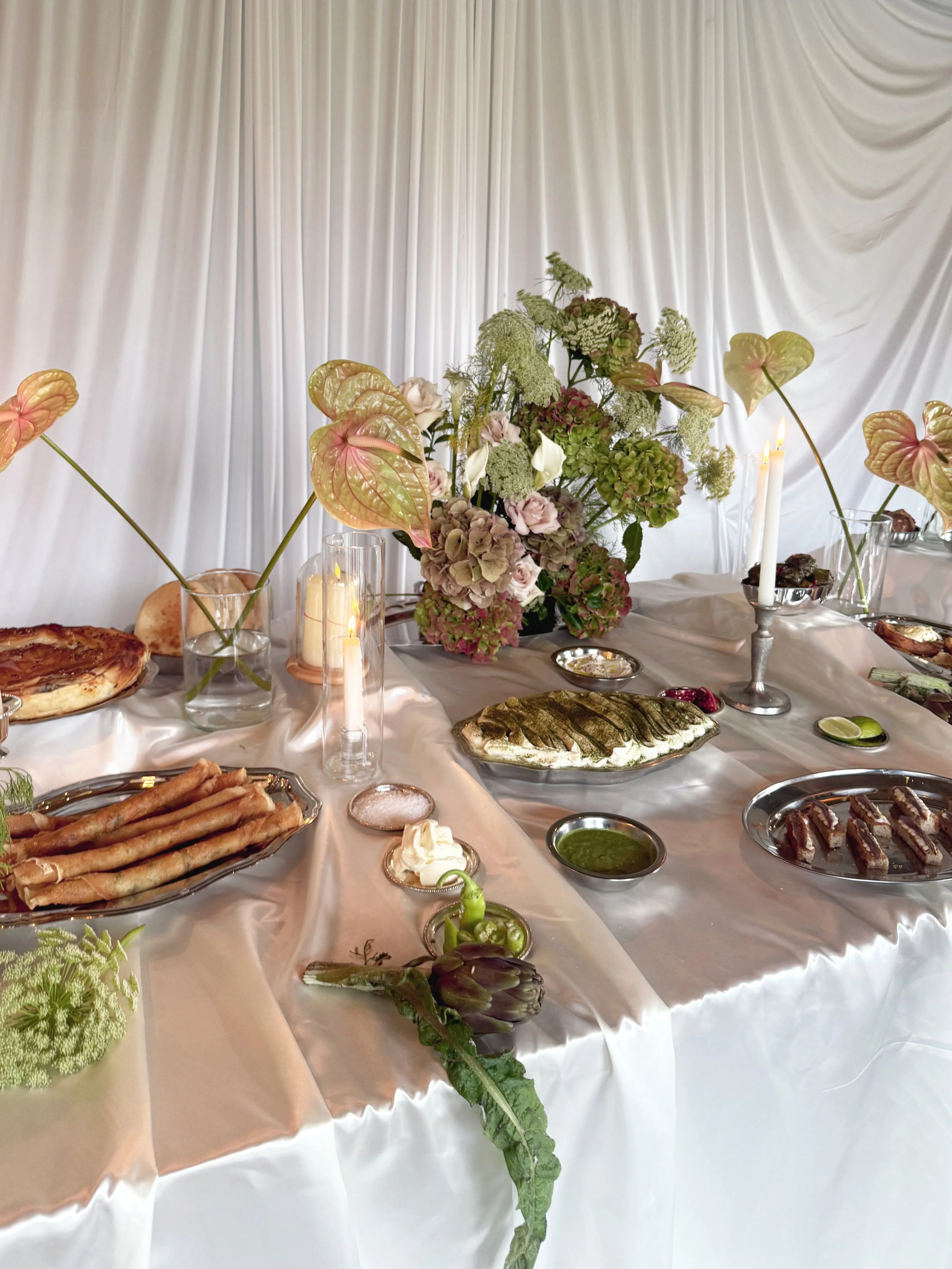 Table avec un arrangement floral composé de fleurs blanches et vertes, servies de bougies allumées, entourée de plats de nourriture comme des tartes, des olives, des sauces, des pâtisseries, et divers mets, devant un fond en rideau blanc.
