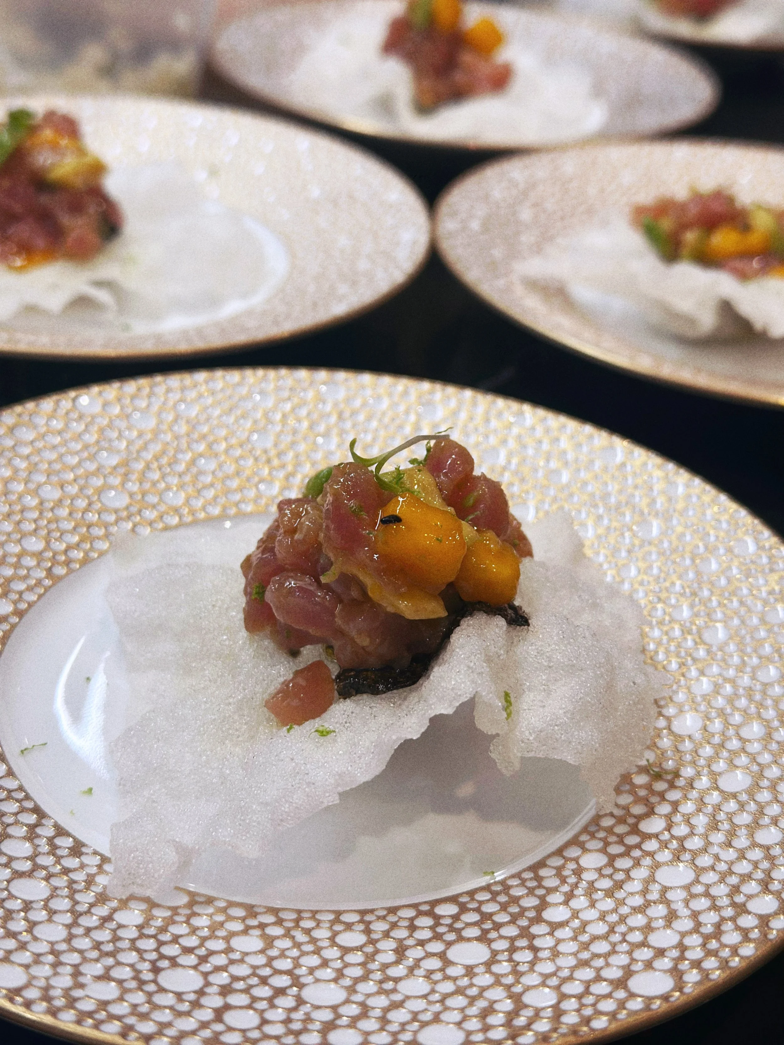 Une assiette avec un plat de tartare de poisson décoré avec des morceaux de fruits ou légumes, présenté sur une assiette blanche avec un motif doré.