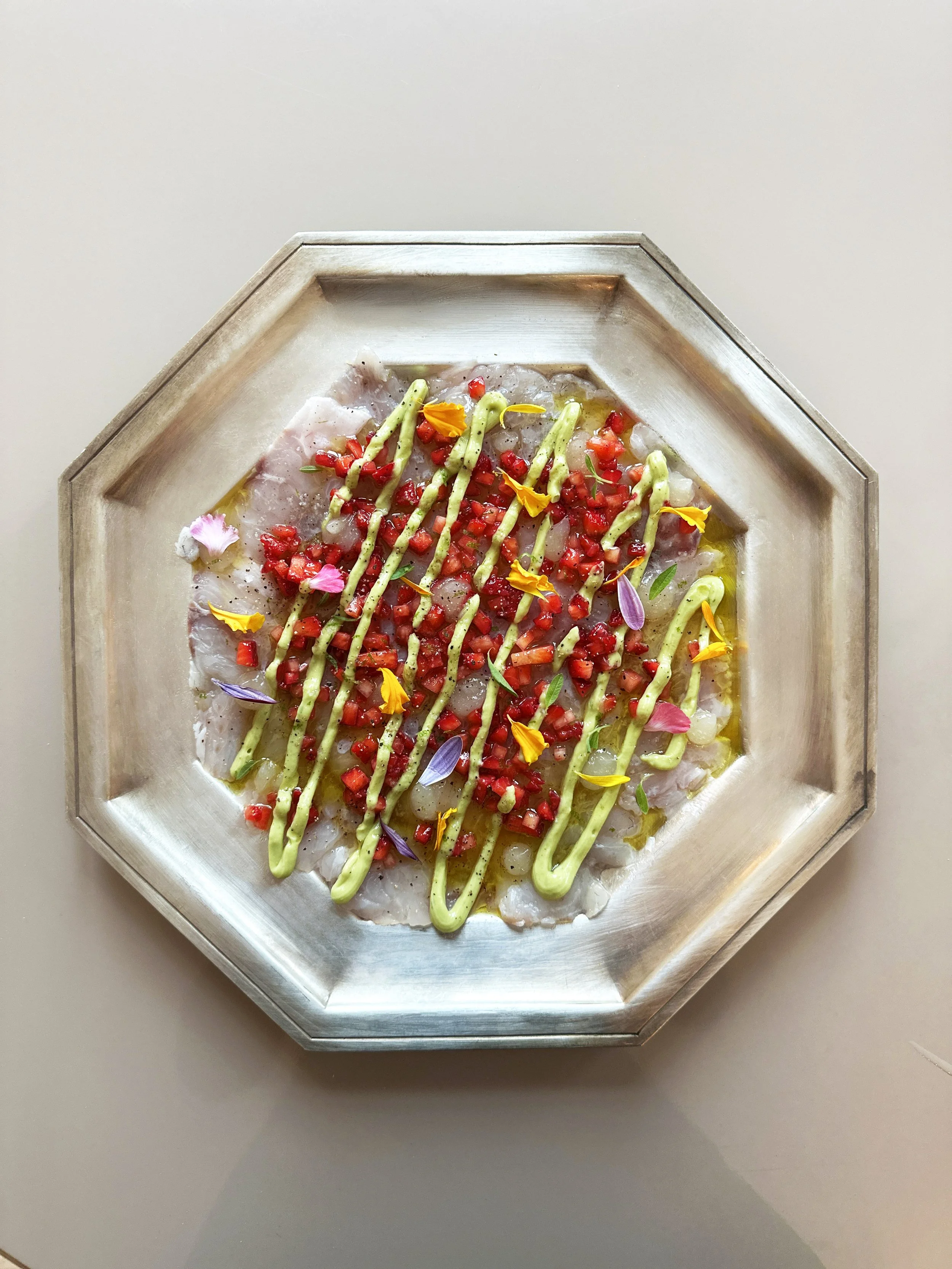 Un plat de poisson cru avec des fraises, de fleurs comestibles en différentes couleurs, avec des lignes de sauce verte à l'avocat, gel citron sur le dessus, présenté dans un plat en métal argenté