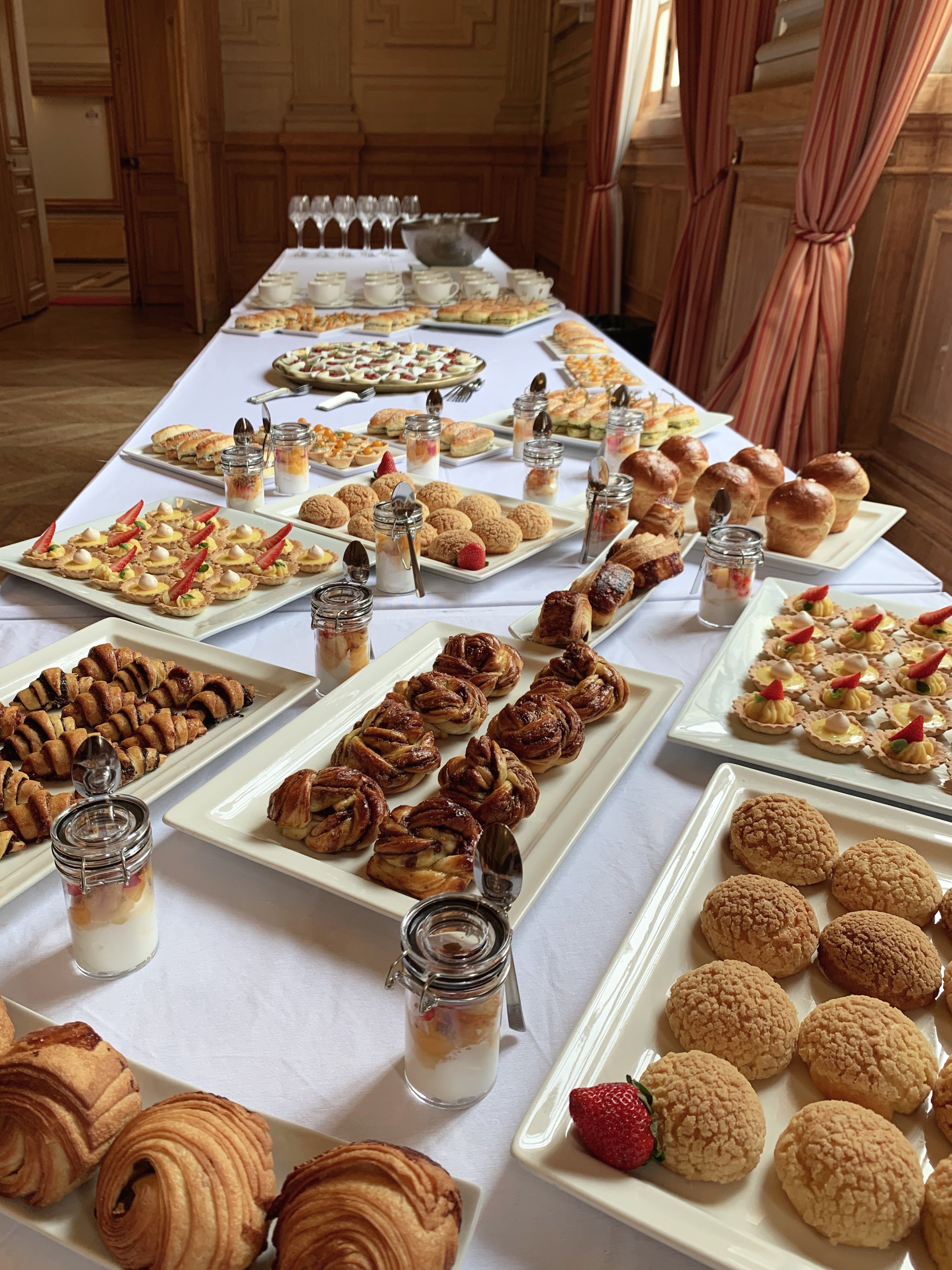 Un buffet sucré avec des biscuits, des pâtisseries, des cupcakes, des tartelettes, des cookies, des choux et des verrines de desserts, posé sur une table avec un fond en bois et des rideaux rouges.